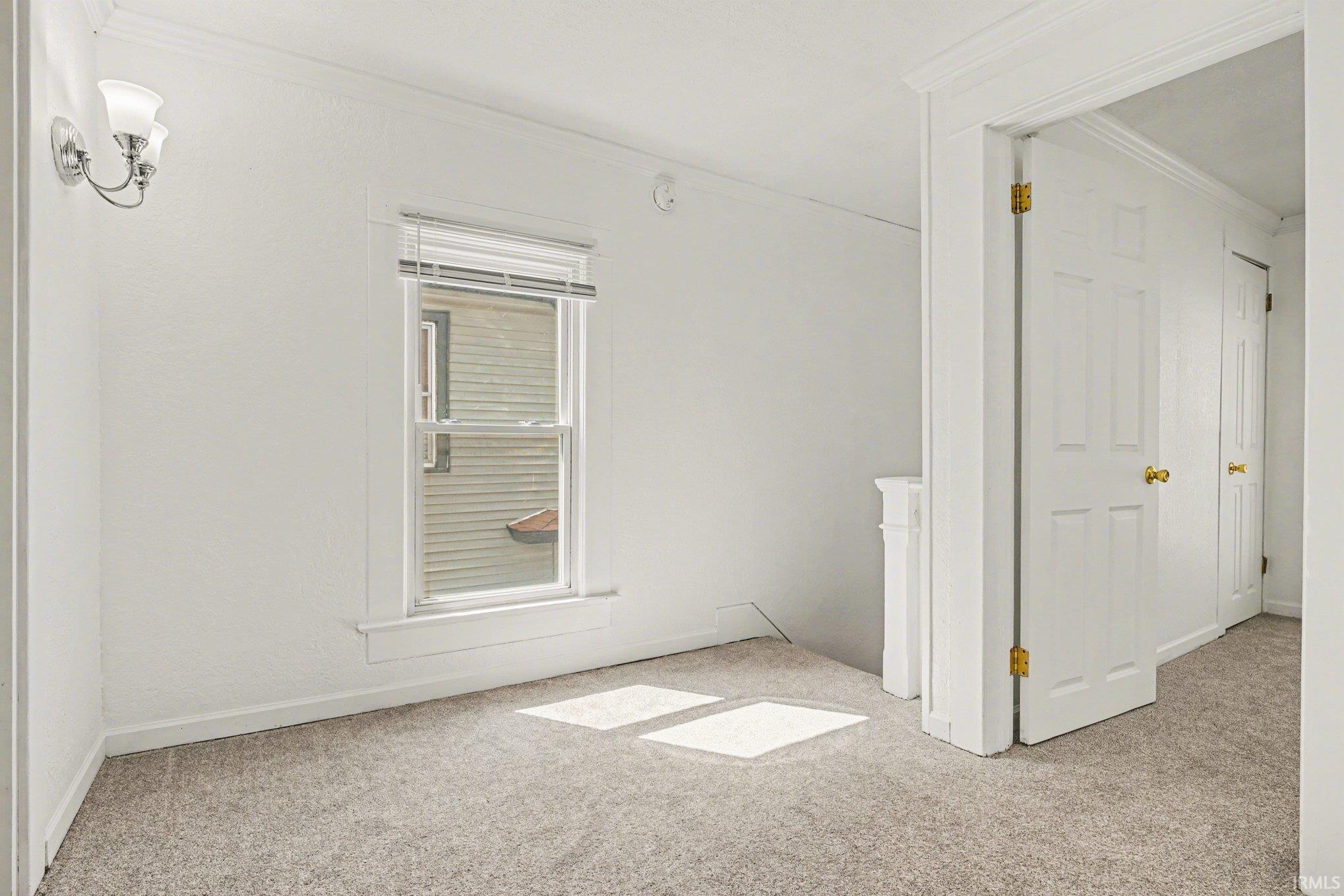 Carpeted room featuring a single window with blinds, a wall-mounted light fixture, a paneled door with a brass-finish knob, and a white architectural element