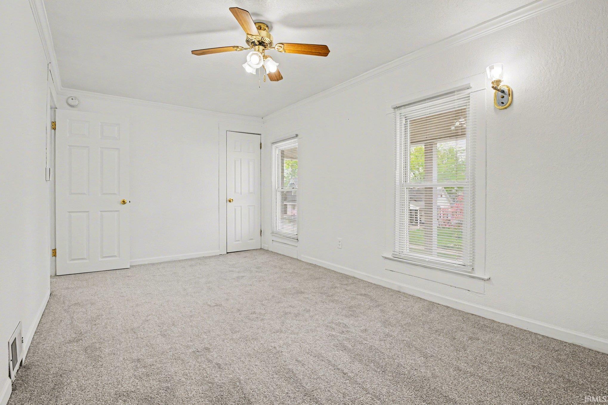 Carpeted interior space featuring a ceiling fan with integrated lighting, two windows with blinds, and white paneled doors with brass-finish hardware