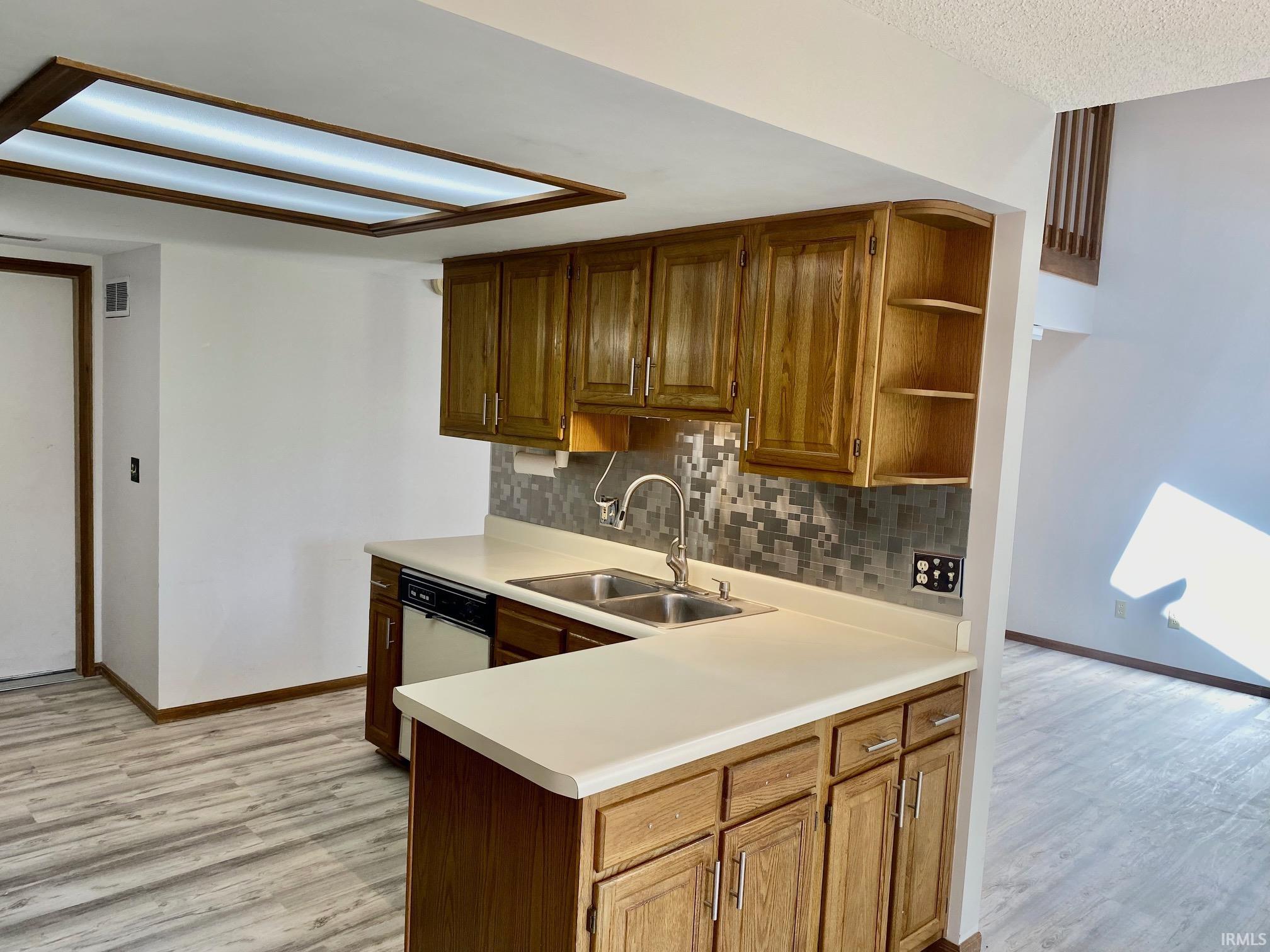 Kitchen featuring wood-finish cabinetry, light-toned countertops, a stainless steel double basin sink with a high-arc faucet, a mosaic tile backsplash, and wood-finish flooring