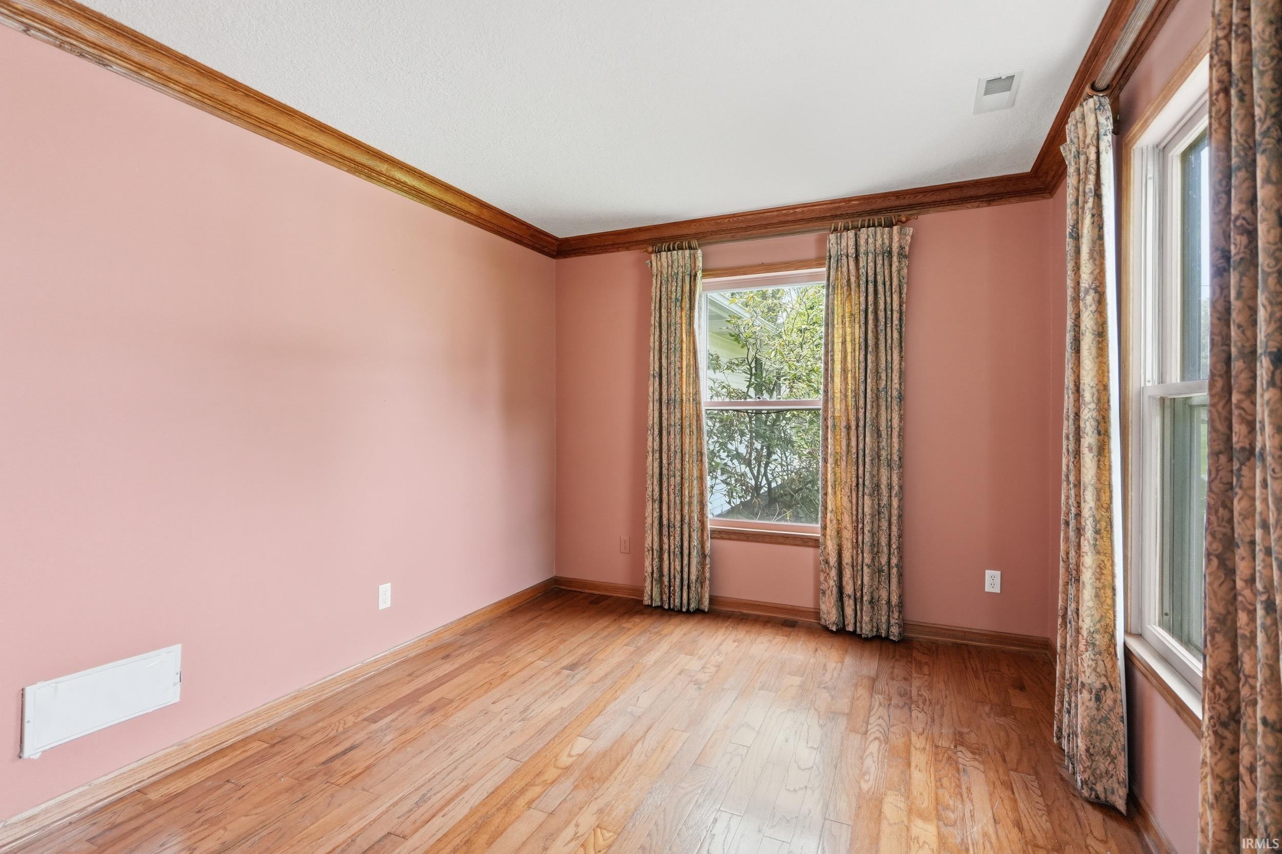 Room featuring wood-finish flooring, two windows with patterned drapes, crown molding, and baseboards