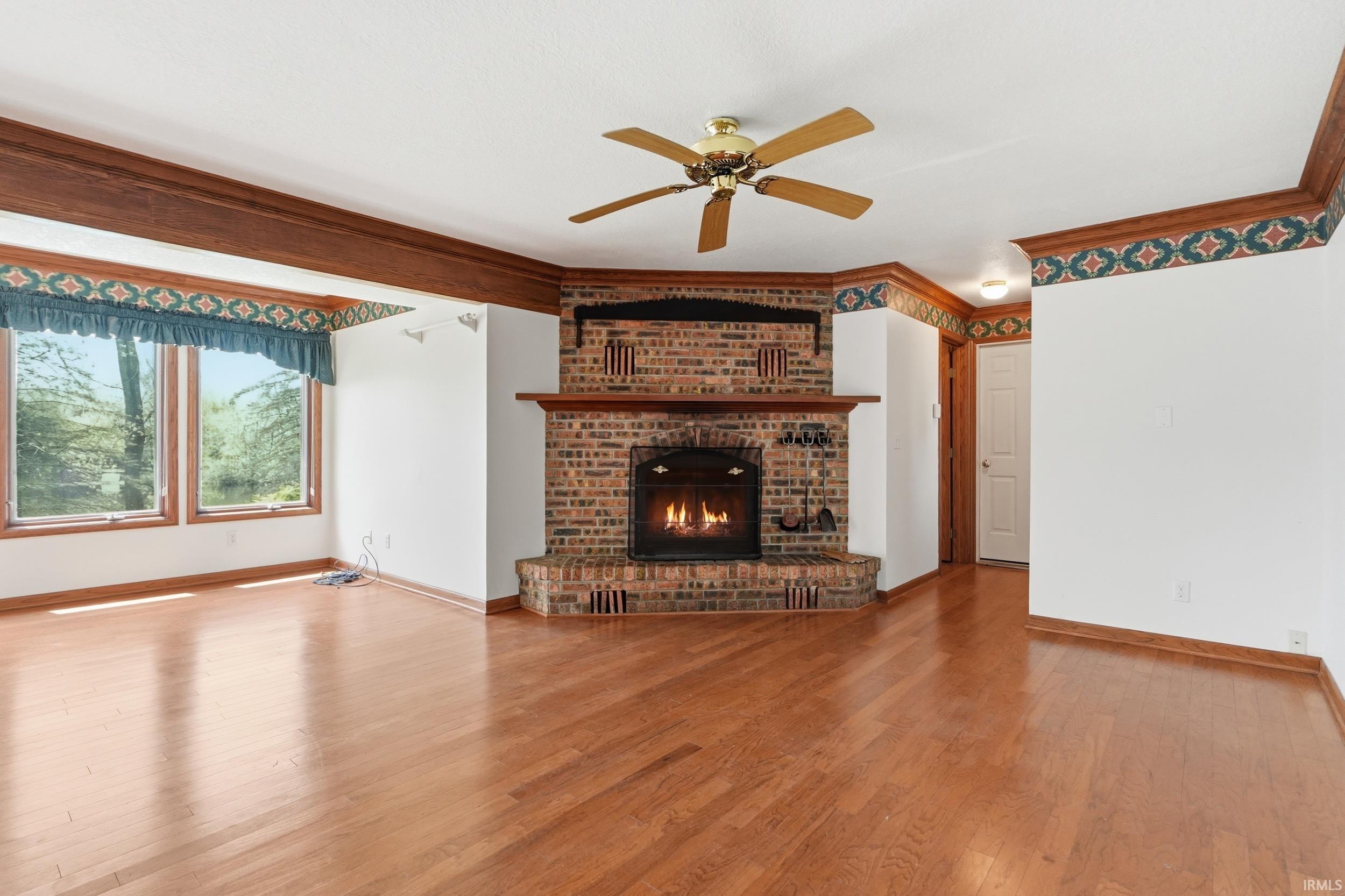 Spacious living area featuring a brick fireplace with a wood mantle, wood-finish flooring, a ceiling fan, and large windows