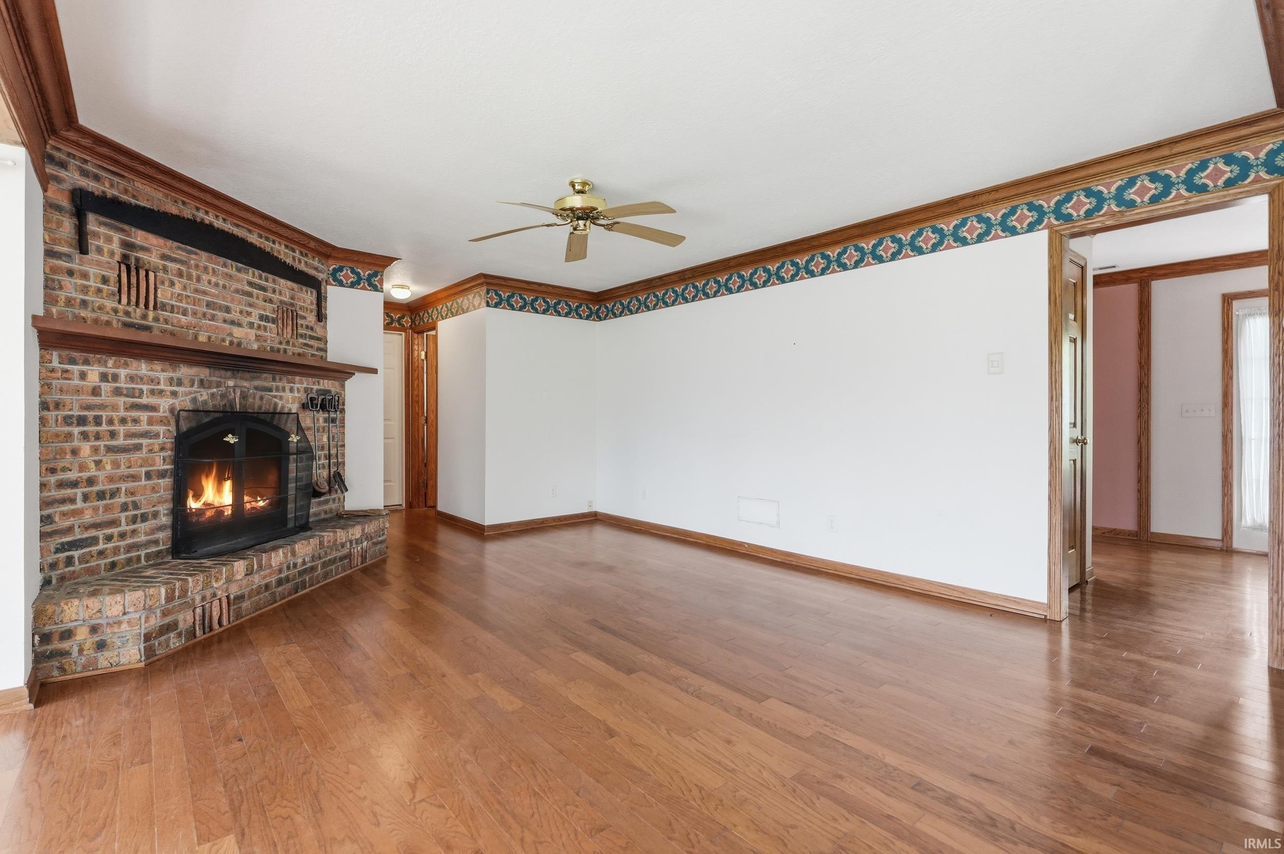 Spacious living area featuring a prominent brick fireplace with a wood mantel, rich wood-finish flooring, and a ceiling fan