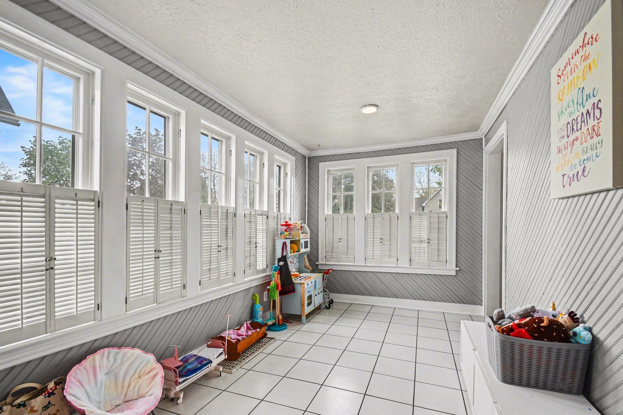 Sunroom featuring multiple windows with plantation shutters, white square tile flooring, beadboard wall paneling, crown molding, and a flush-mount ceiling light