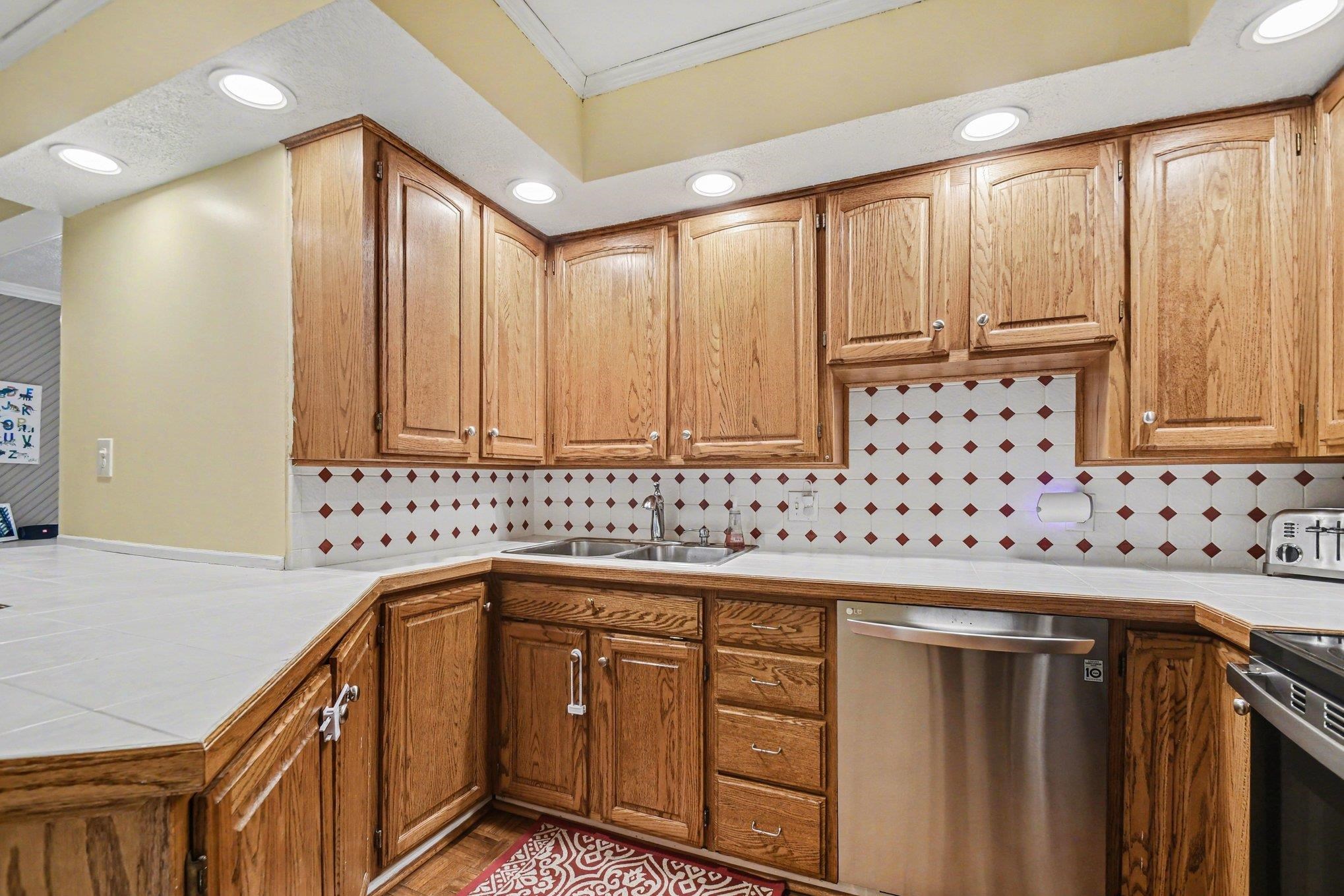 Kitchen featuring wood-finish cabinetry, tile countertops, a stainless steel dishwasher, an integrated dual basin sink, and recessed lighting