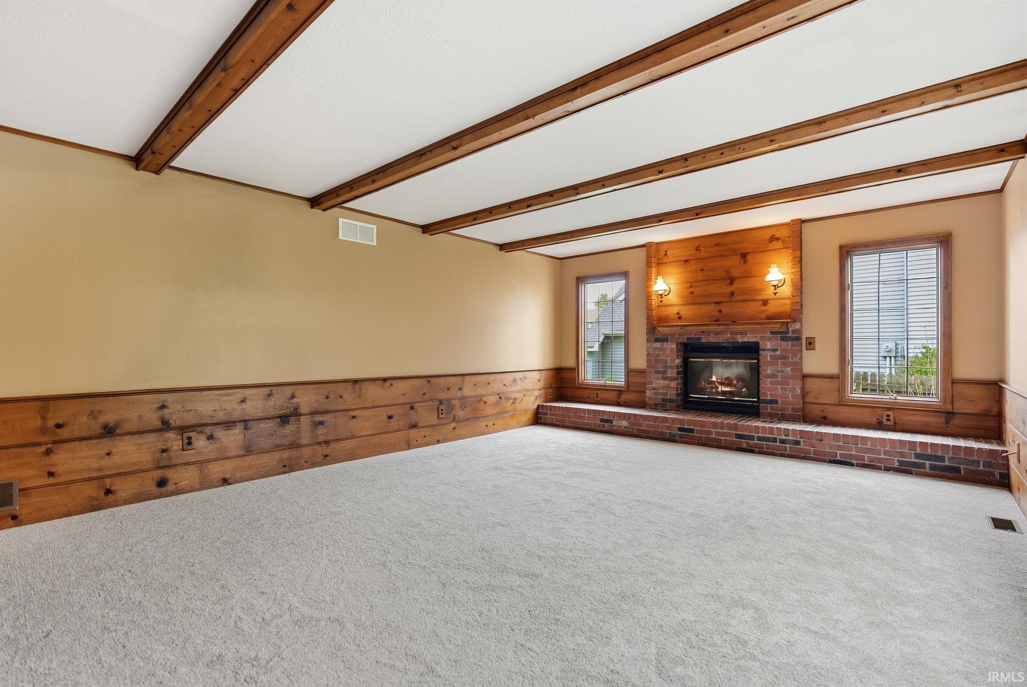 Spacious room featuring a brick fireplace with a wood mantle, wood beam ceiling, wood-paneled wainscoting, and light-toned carpeting
