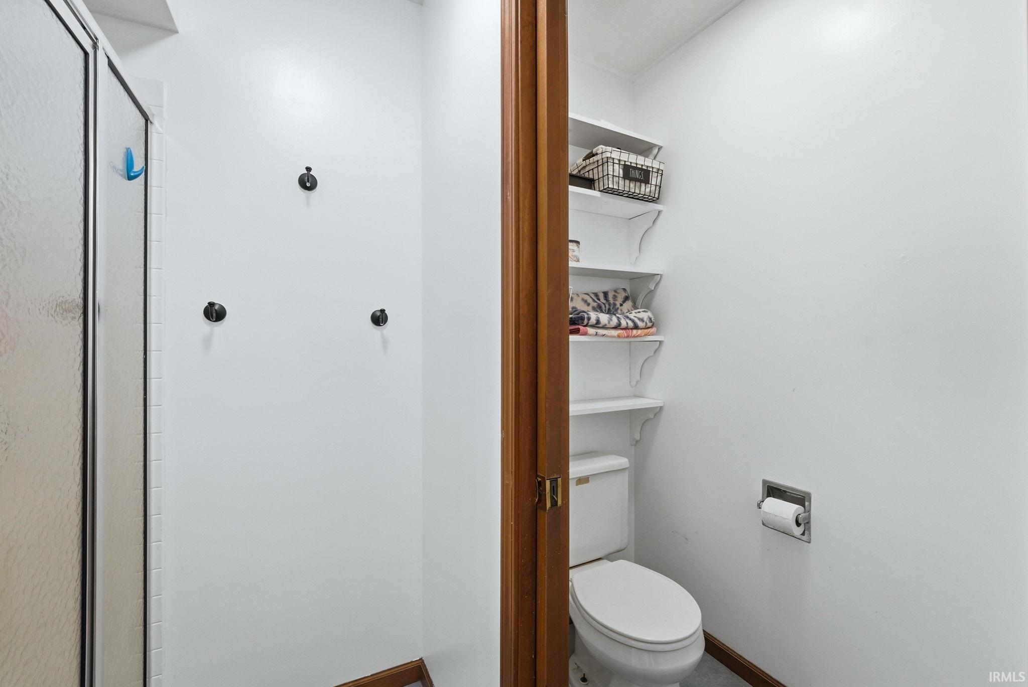Shower enclosure with frosted glass door, adjacent to a water closet featuring built-in shelving, a standard toilet, and a wall-mounted toilet paper holder