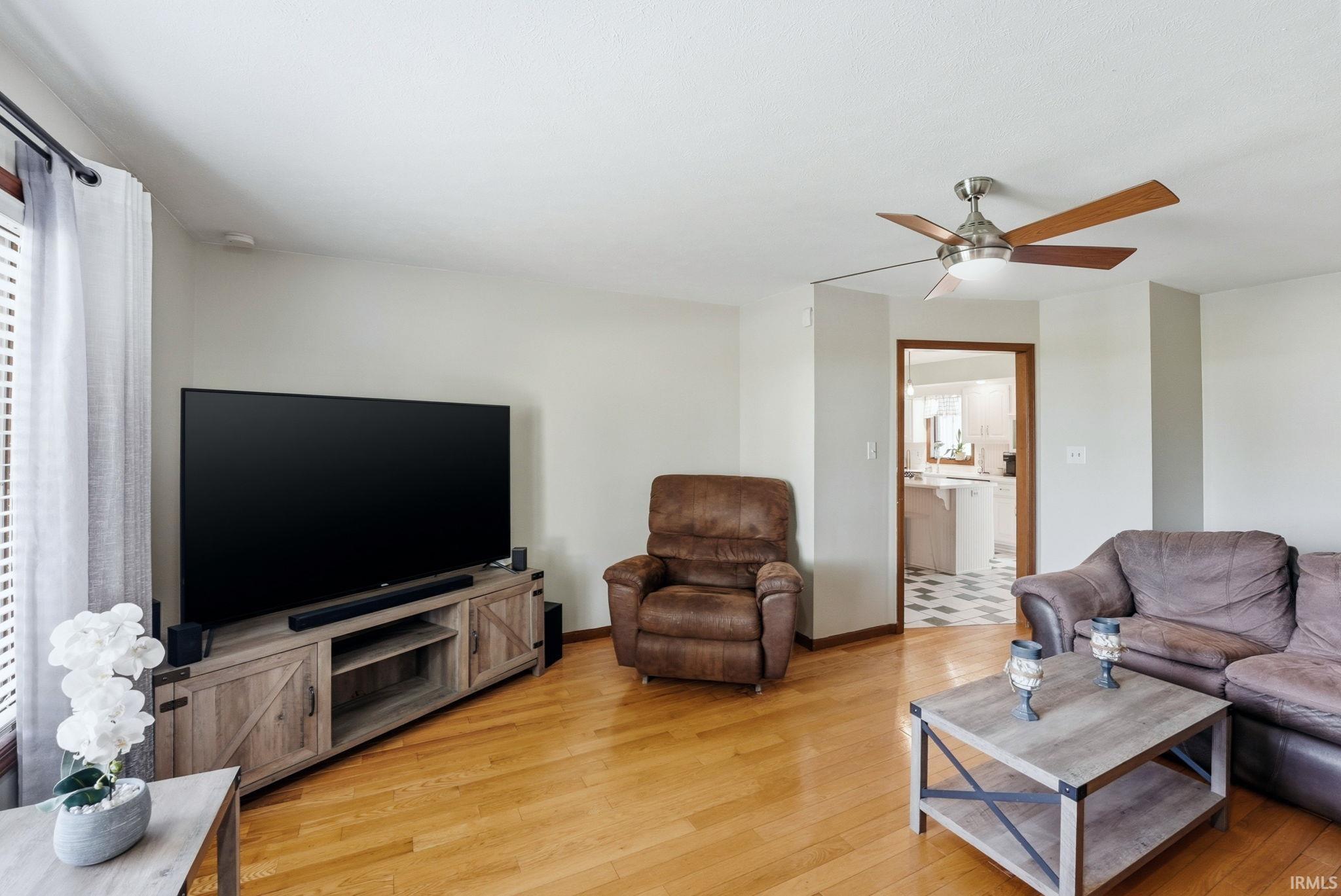 Spacious living area featuring wood-finish flooring, a ceiling fan with wood-tone blades, and white painted walls