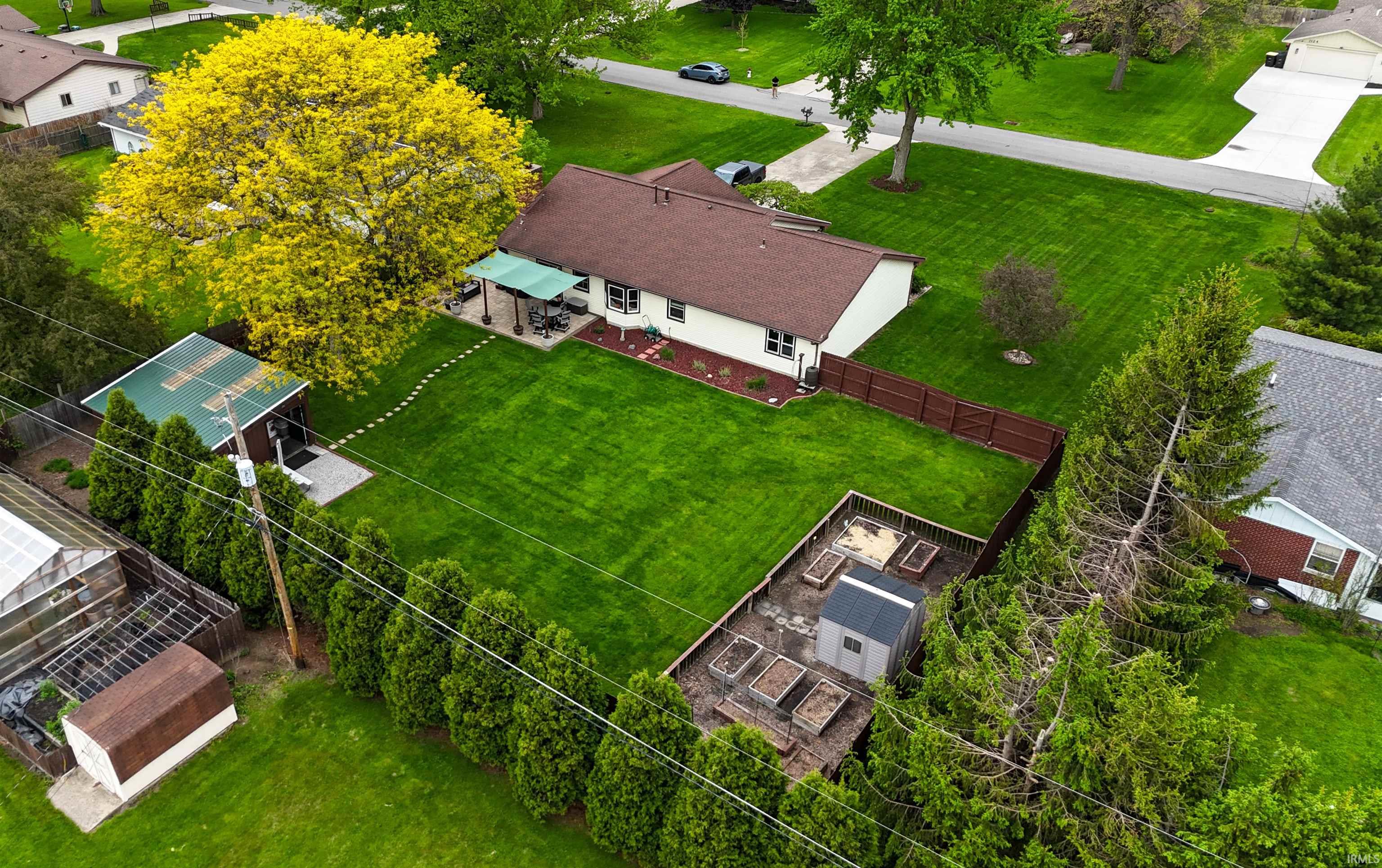 Expansive property featuring a detached shed with a green metal roof, a patio with an overhead awning, a dedicated garden area with raised beds, mature perimeter plantings, and a brown privacy fence