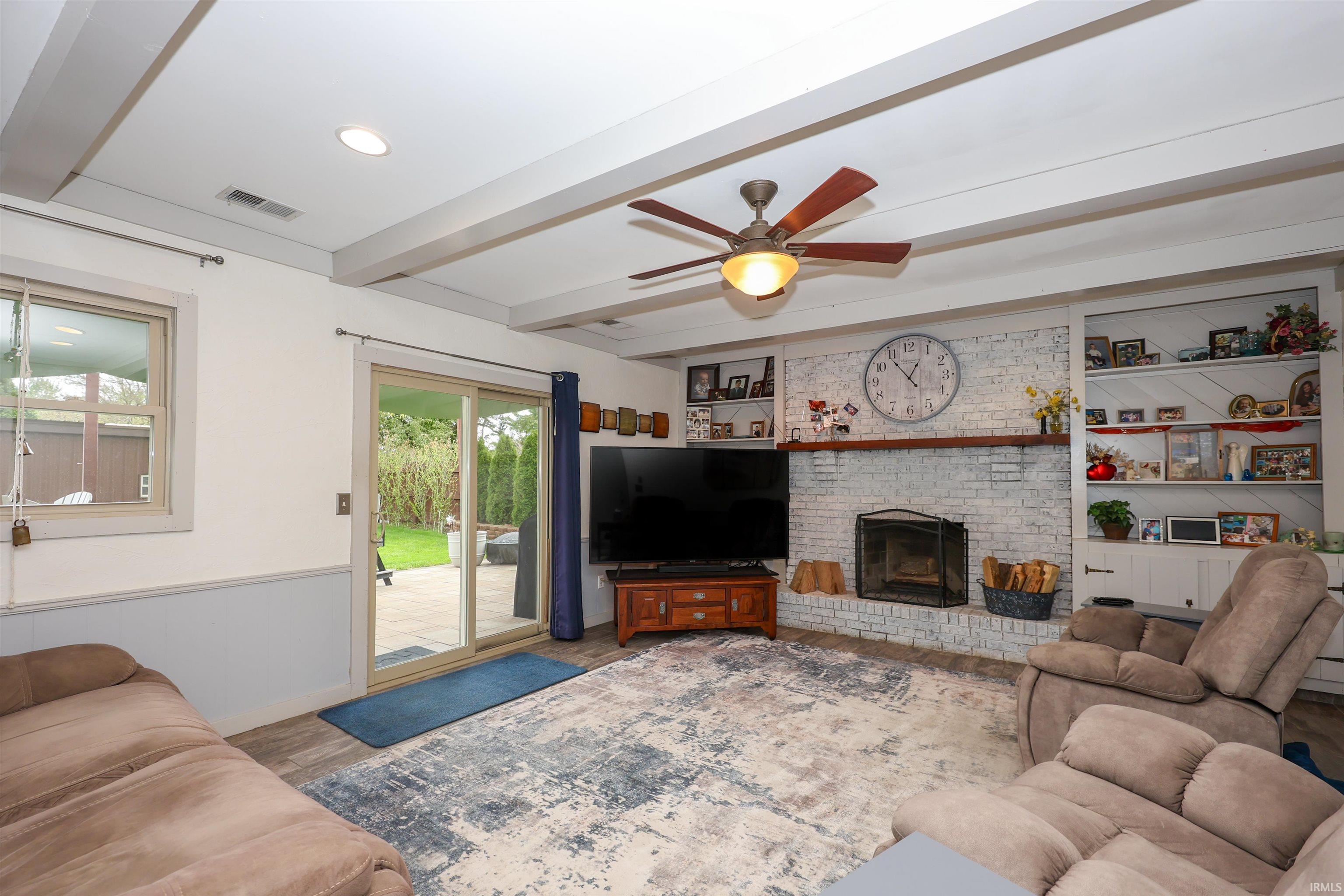 Living space featuring a whitewashed brick fireplace with a wood mantle, built-in shelving, wood-finish flooring, and exposed ceiling beams