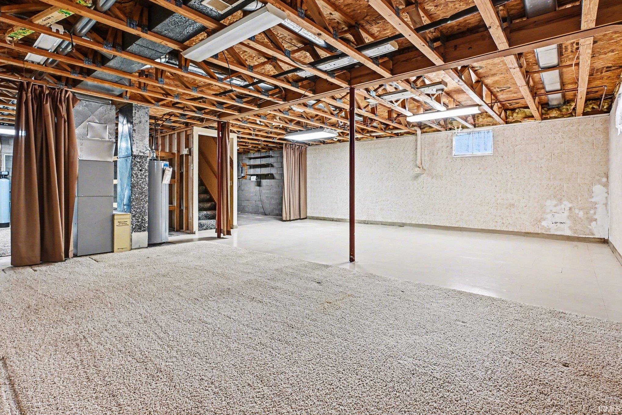 Unfinished basement featuring exposed wood joists and structural beams, offering an expansive area with both carpeted and concrete flooring sections