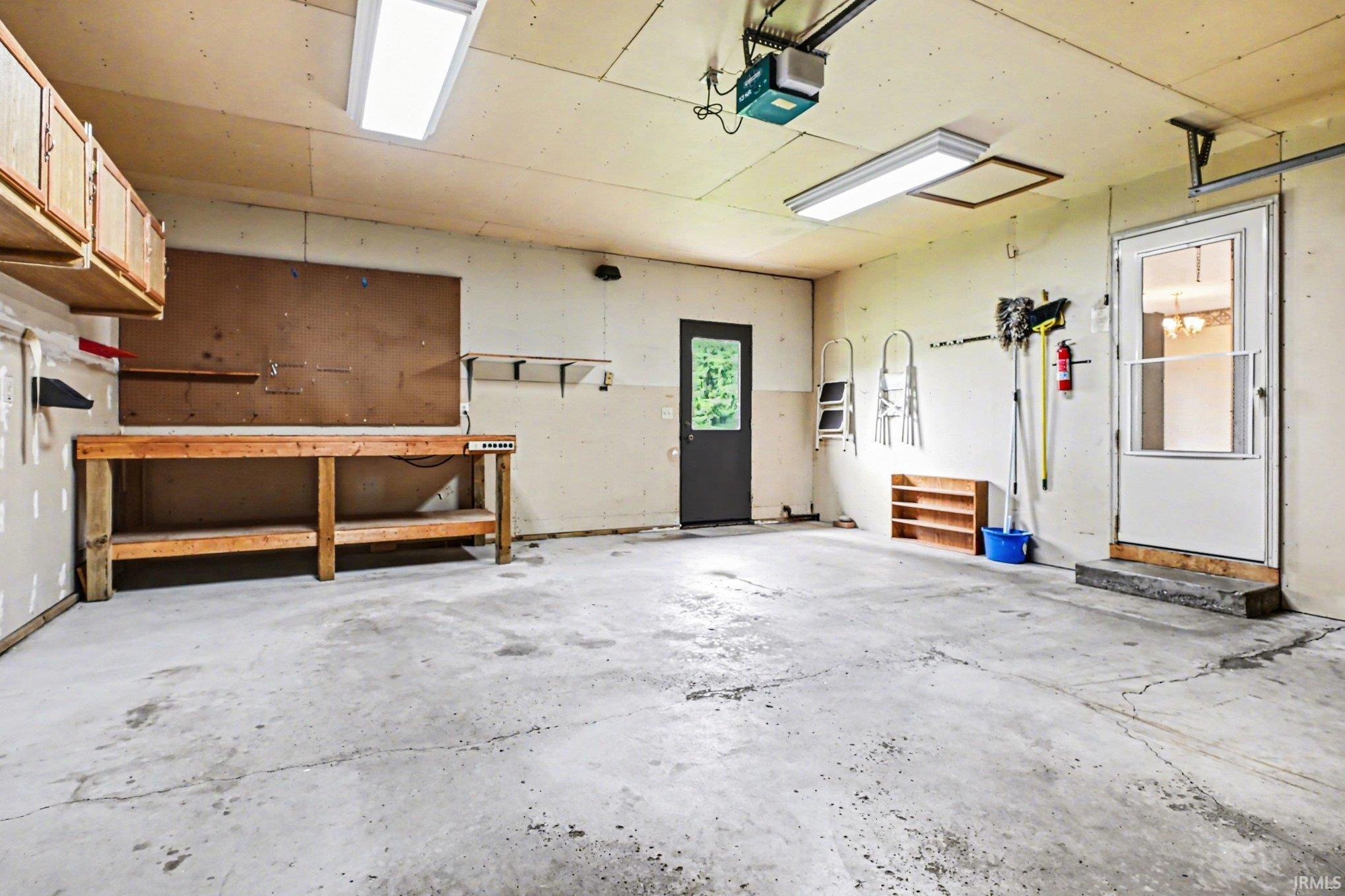 Concrete floor garage with two permanent entry doors, overhead fluorescent lighting, and a built-in wooden workbench with overhead cabinetry