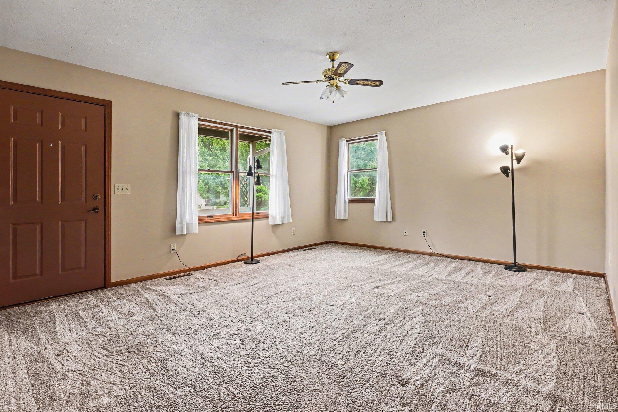 Spacious room featuring neutral carpeting, a panel door, dual windows with wood trim, and a ceiling fan with integrated lighting