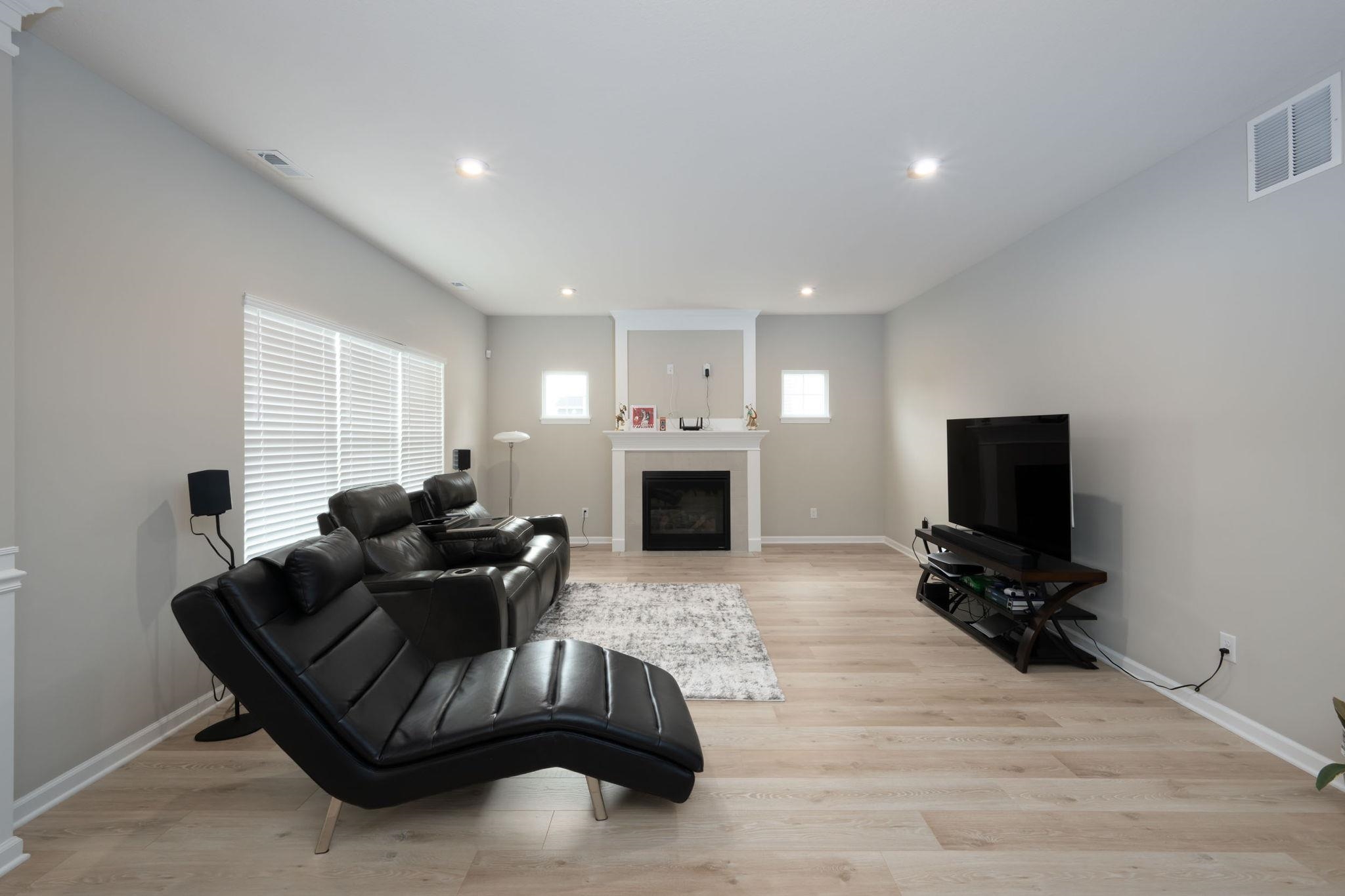 Spacious living area featuring light wood-finish flooring, a white fireplace with mantle, recessed lighting, and ample natural light from multiple windows