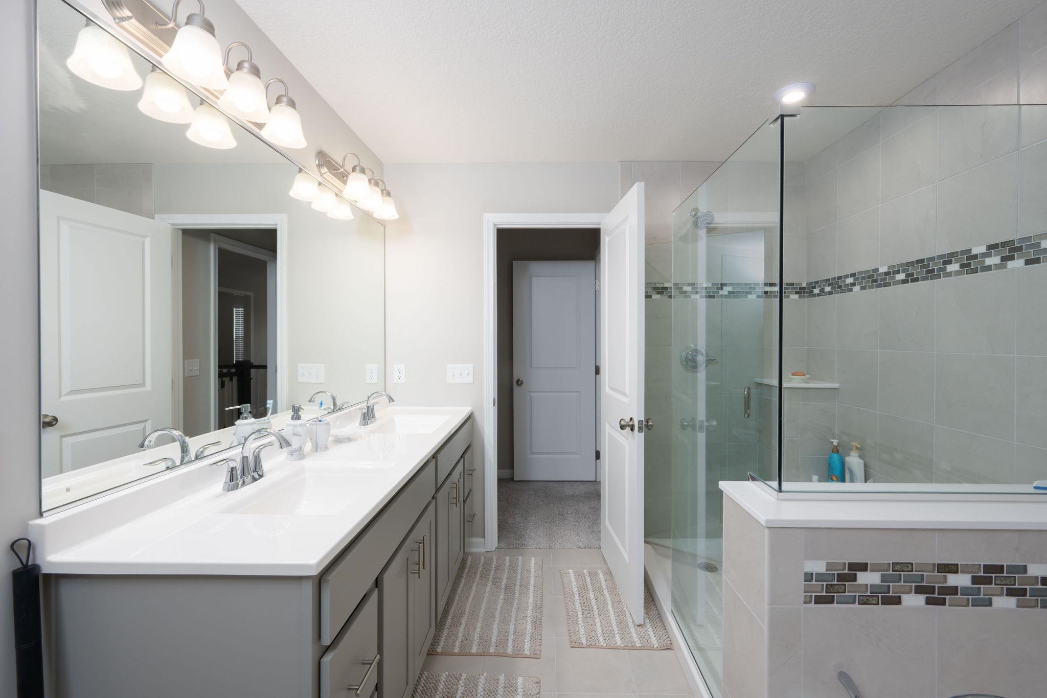 Spacious bathroom featuring a dual vanity with white countertops, chrome fixtures, a large frameless mirror, and a walk-in shower with a mosaic tile accent band