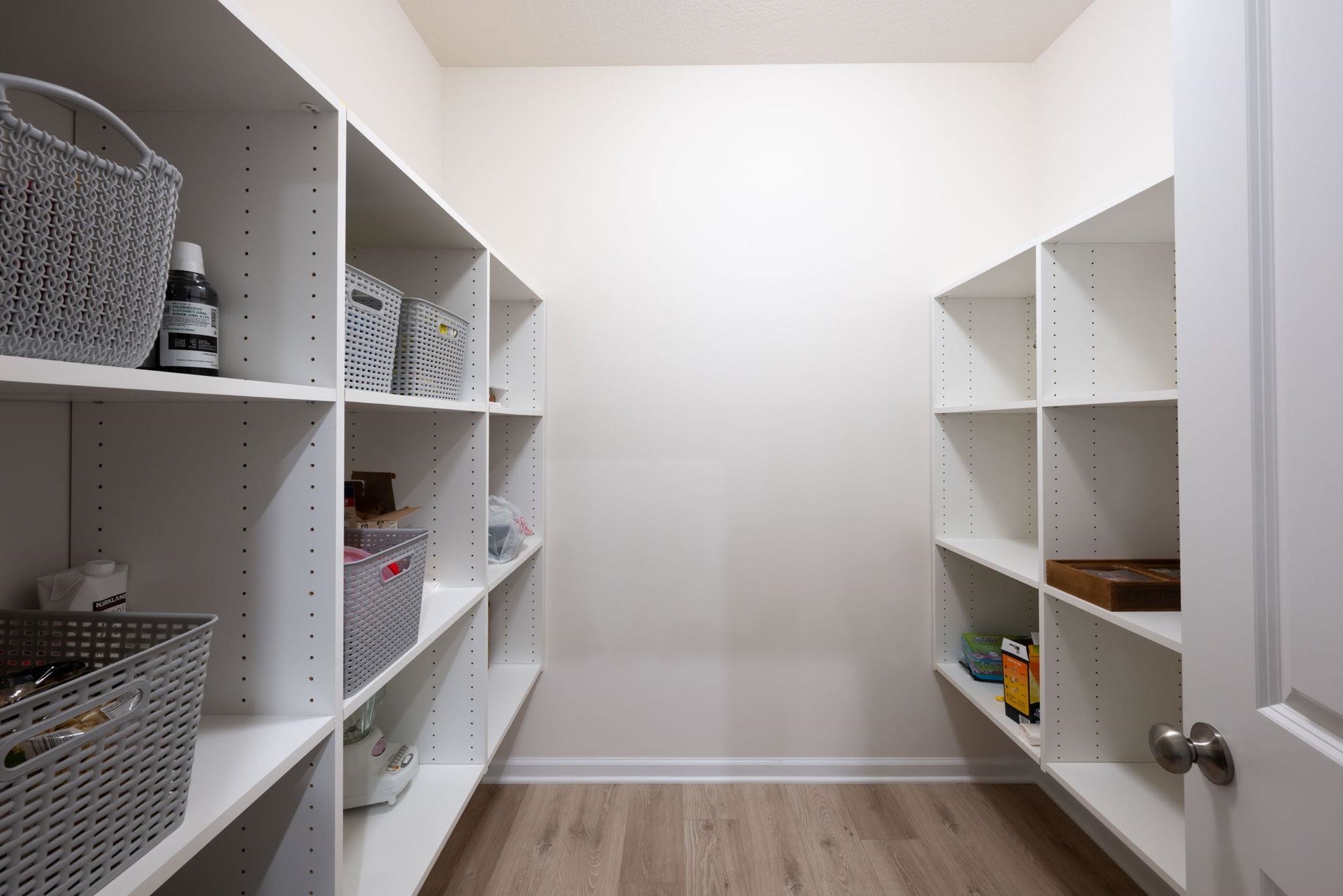 Spacious walk-in pantry featuring white shelving units on opposing walls, wood-finish flooring, and white baseboards