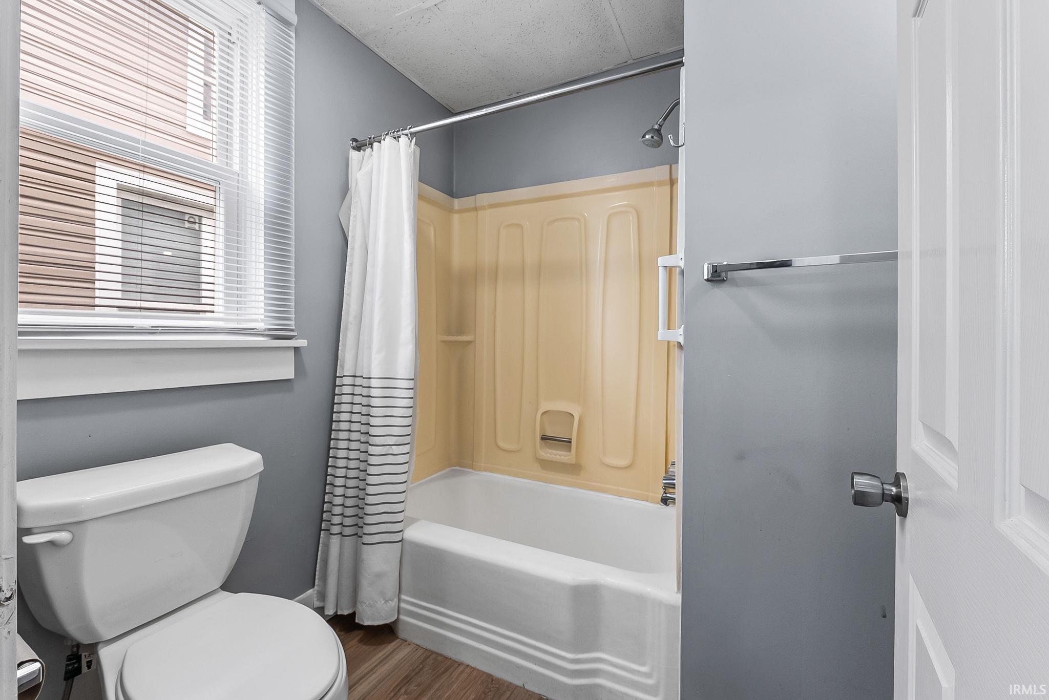 Full bathroom with a white porcelain toilet, bathtub with an integrated shower, and a window with blinds