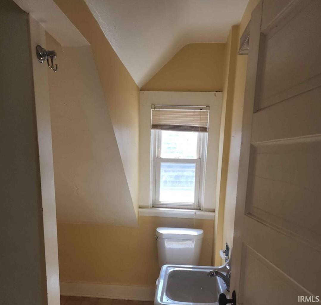 Compact bathroom with sloped ceiling, featuring a window with blinds, a white toilet, and a wall-mounted sink with chrome fixtures
