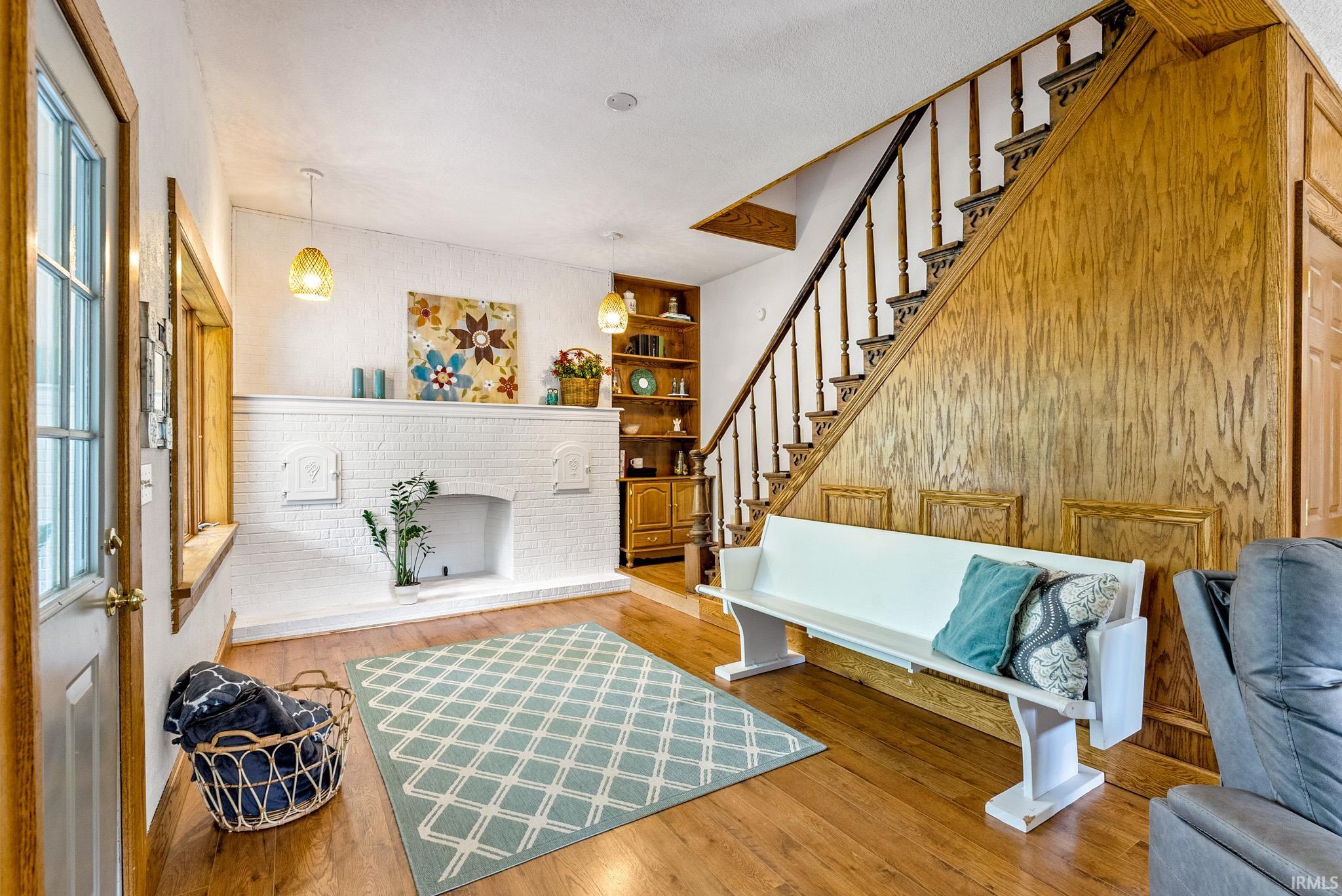 Open living space featuring wood-finish flooring, a white painted brick fireplace with hearth, built-in shelving, and a prominent staircase with wood paneling and balusters