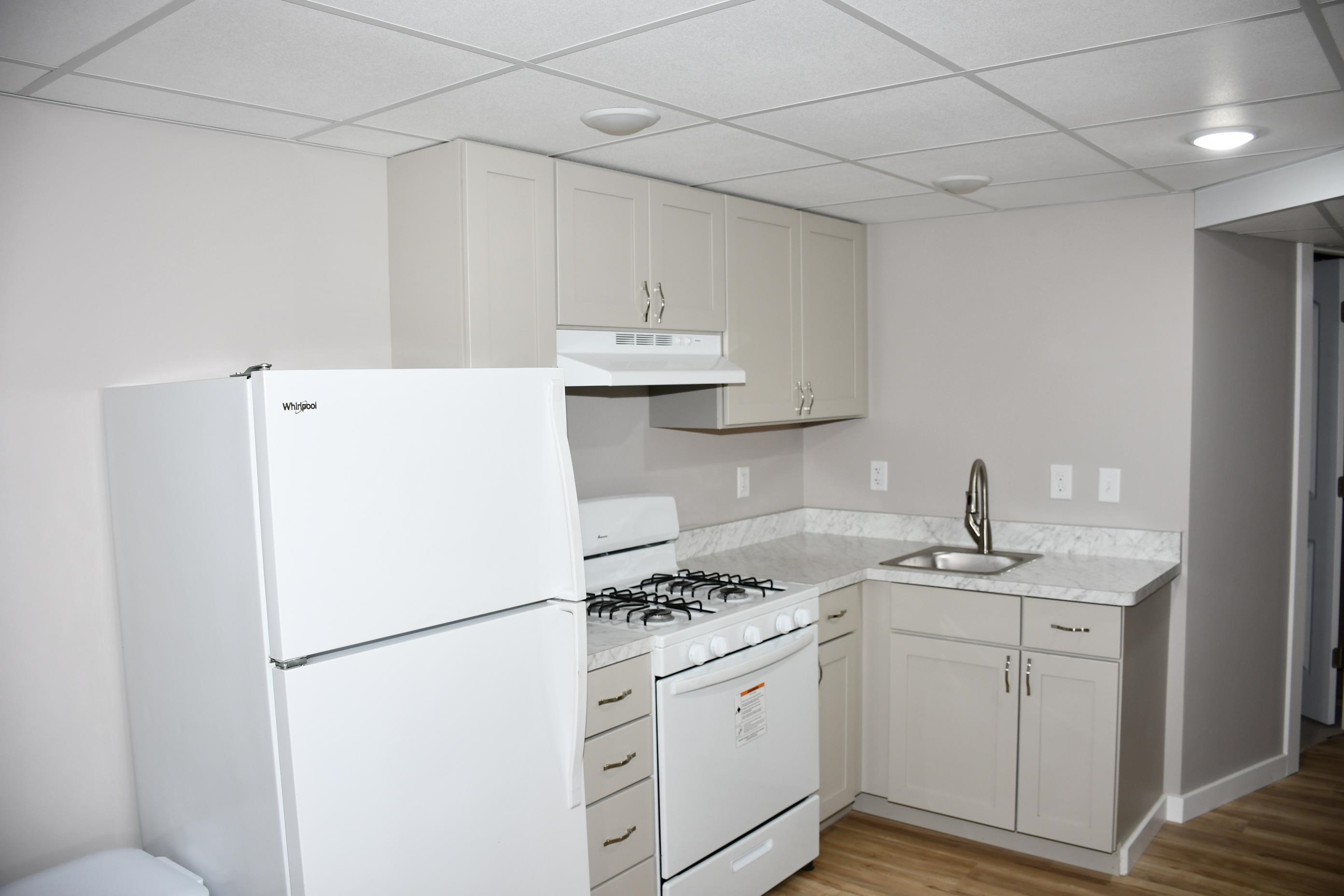 Compact kitchen featuring light gray cabinetry, a white refrigerator, a white gas range with an overhead vent, a stainless steel sink with a gooseneck faucet, and light-toned countertops