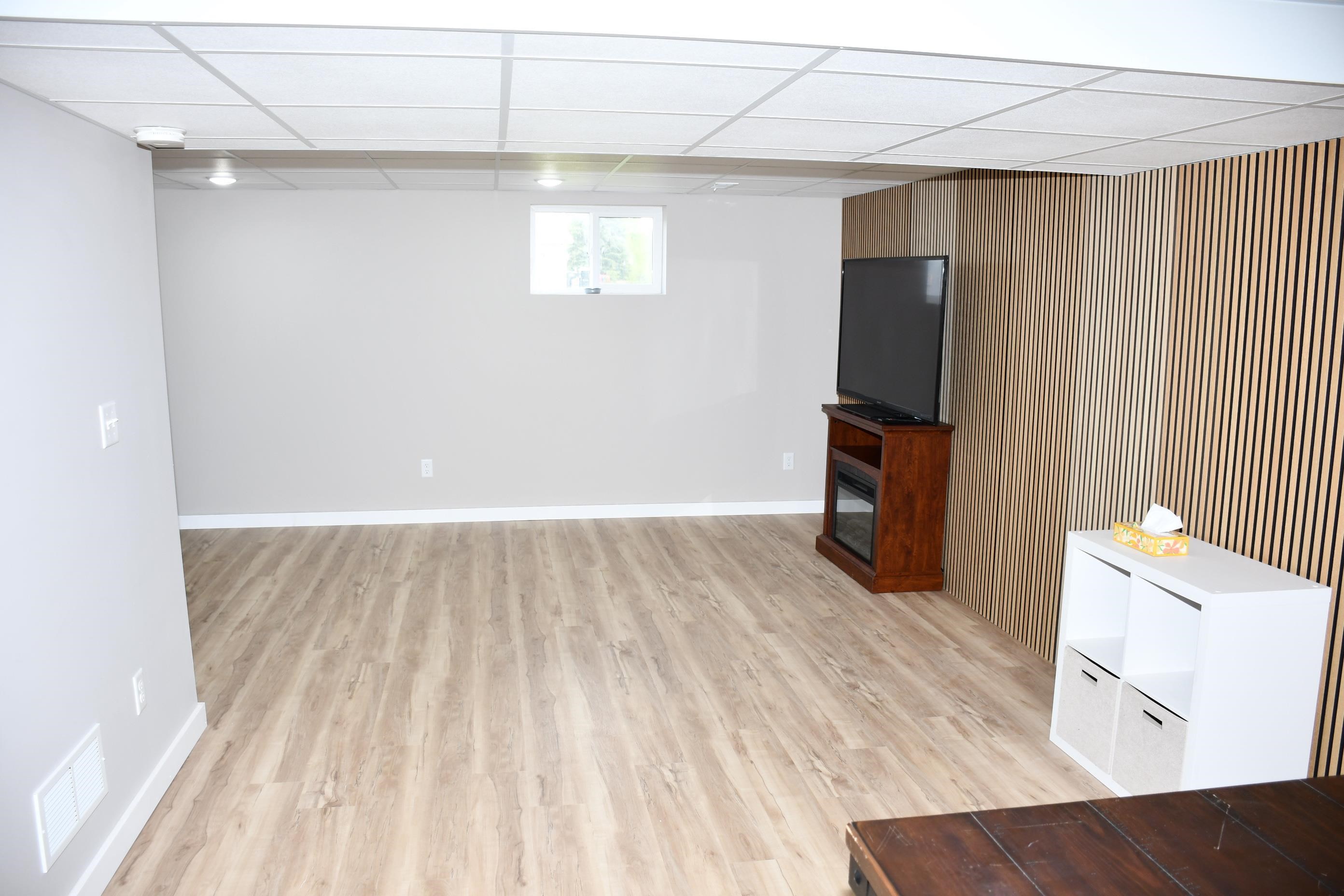 Spacious room featuring light wood-finish flooring, light gray walls, a drop ceiling with recessed lighting, and a horizontal slat wood-paneled accent wall