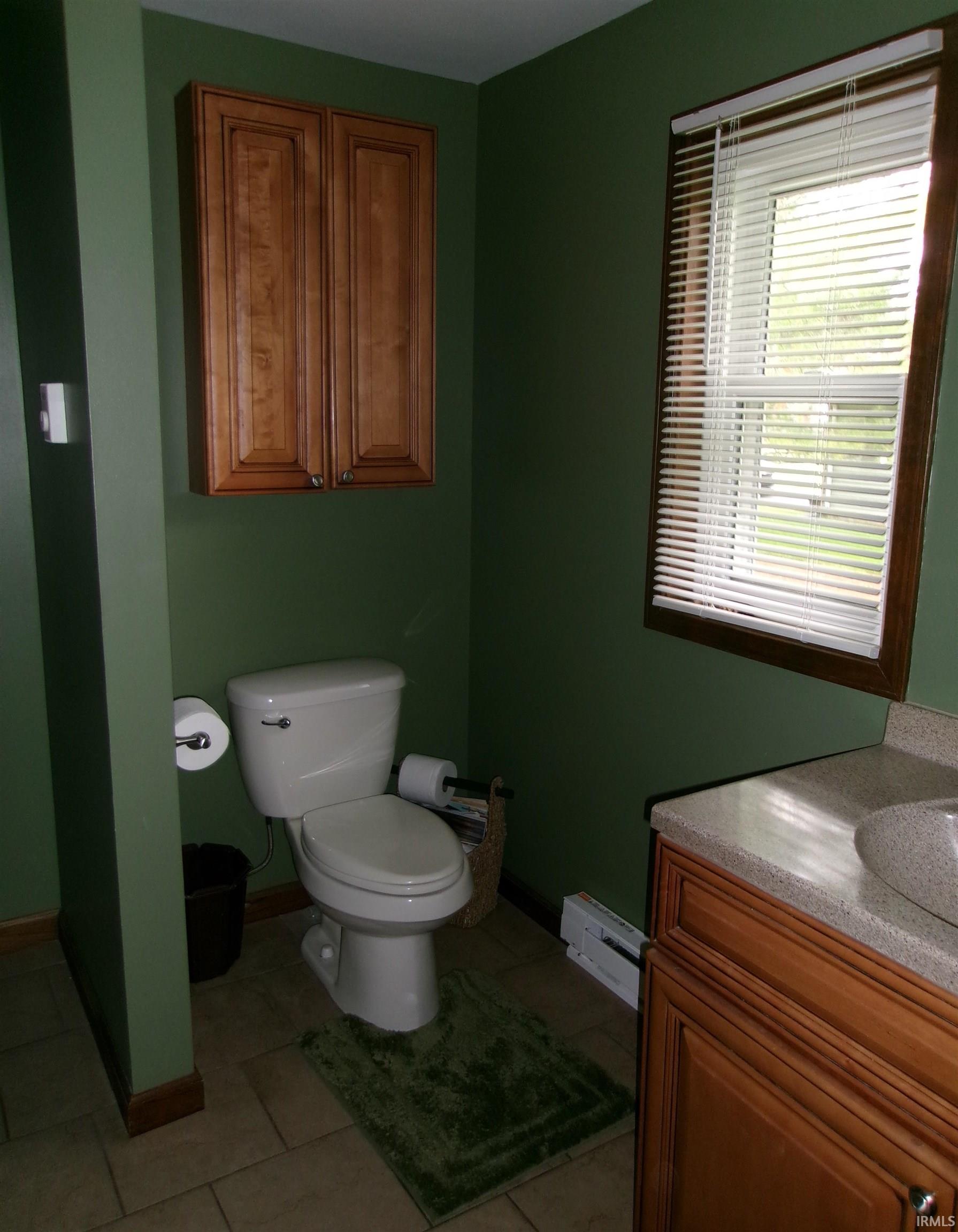 Bathroom featuring a white toilet, wall-mounted wood-finish cabinetry, a window with blinds, a vanity with an integrated sink and light-colored countertop, and tiled flooring