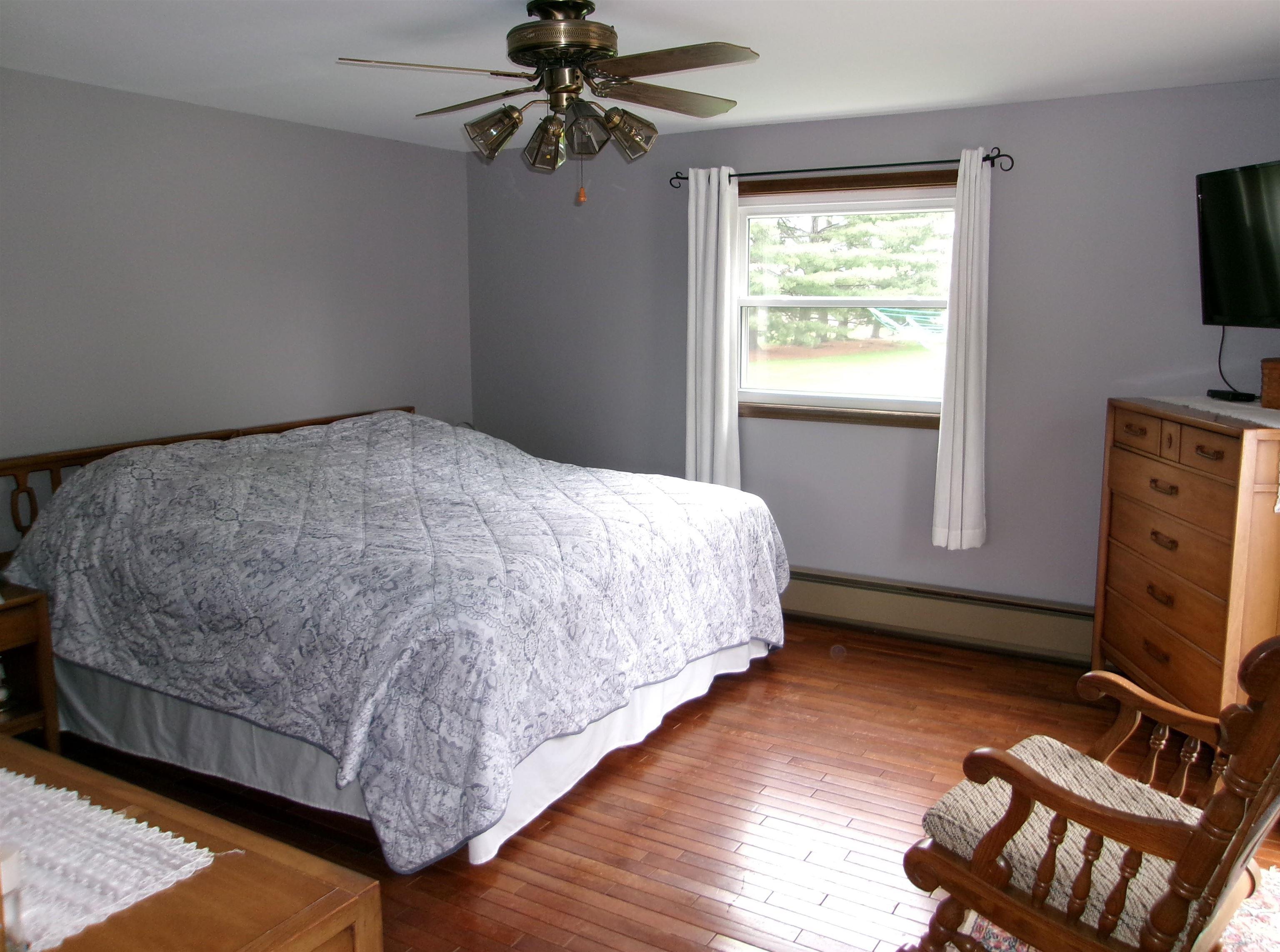 Hardwood flooring throughout this room features a large window with white trim, a ceiling fan with integrated lighting, and neutral wall paint