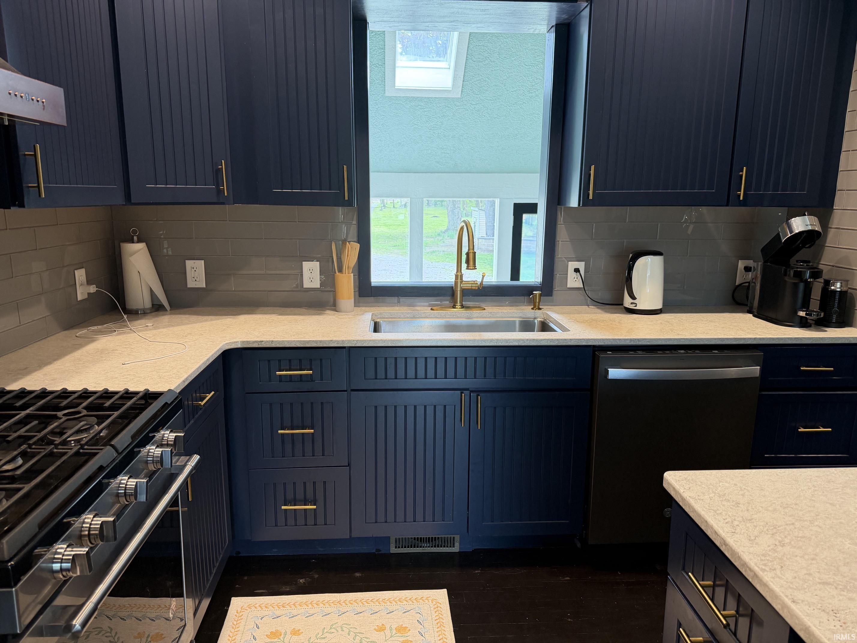 Navy blue cabinetry with gold-tone hardware, light-toned countertops, a stainless steel sink with a gold-tone faucet, a gray subway tile backsplash, and dark wood-finish flooring