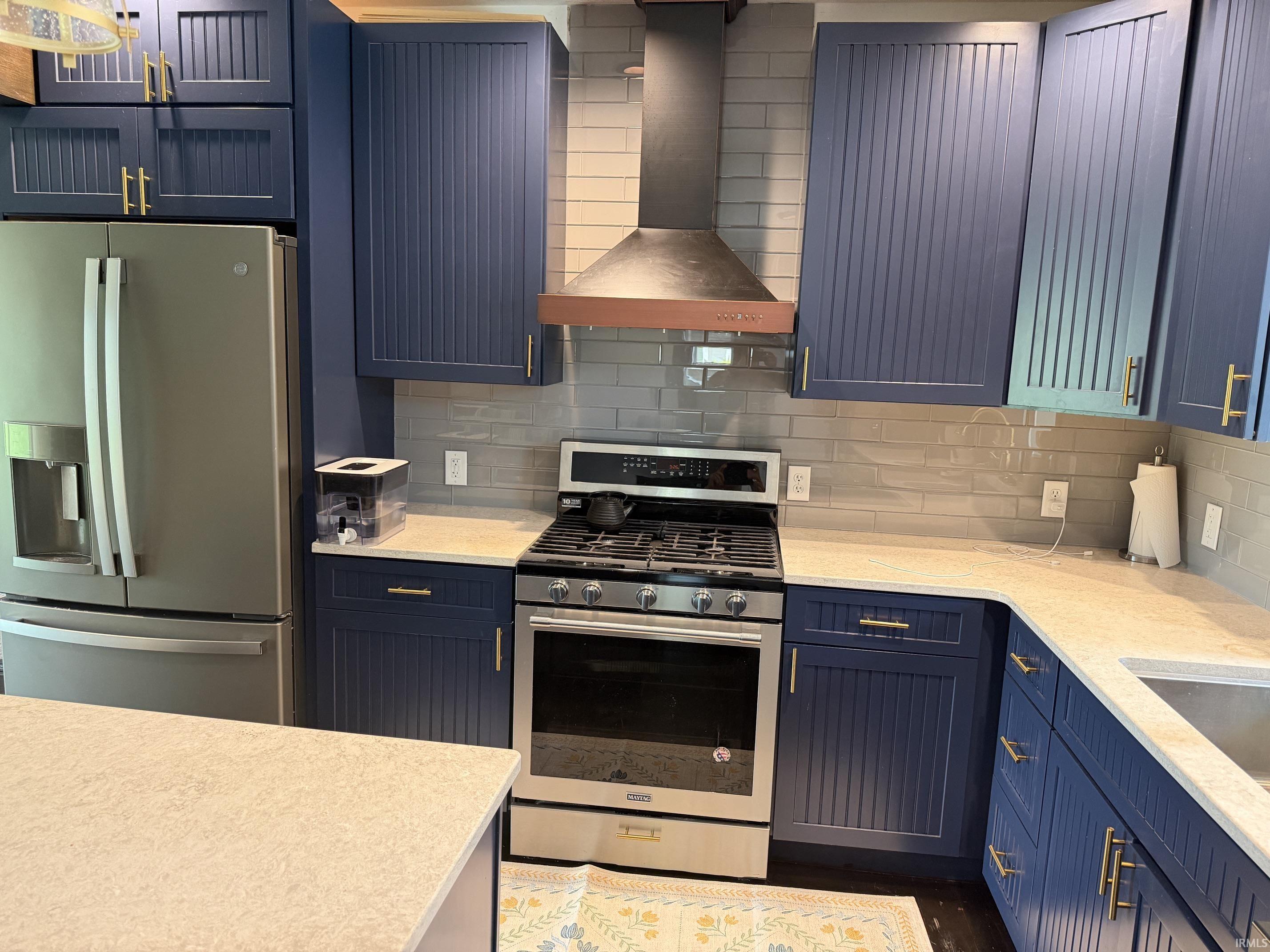 Kitchen featuring dark blue cabinetry with gold-tone hardware, a stainless steel gas range with coordinating hood, a large stainless steel refrigerator, light-toned countertops, and a subway tile backsplash