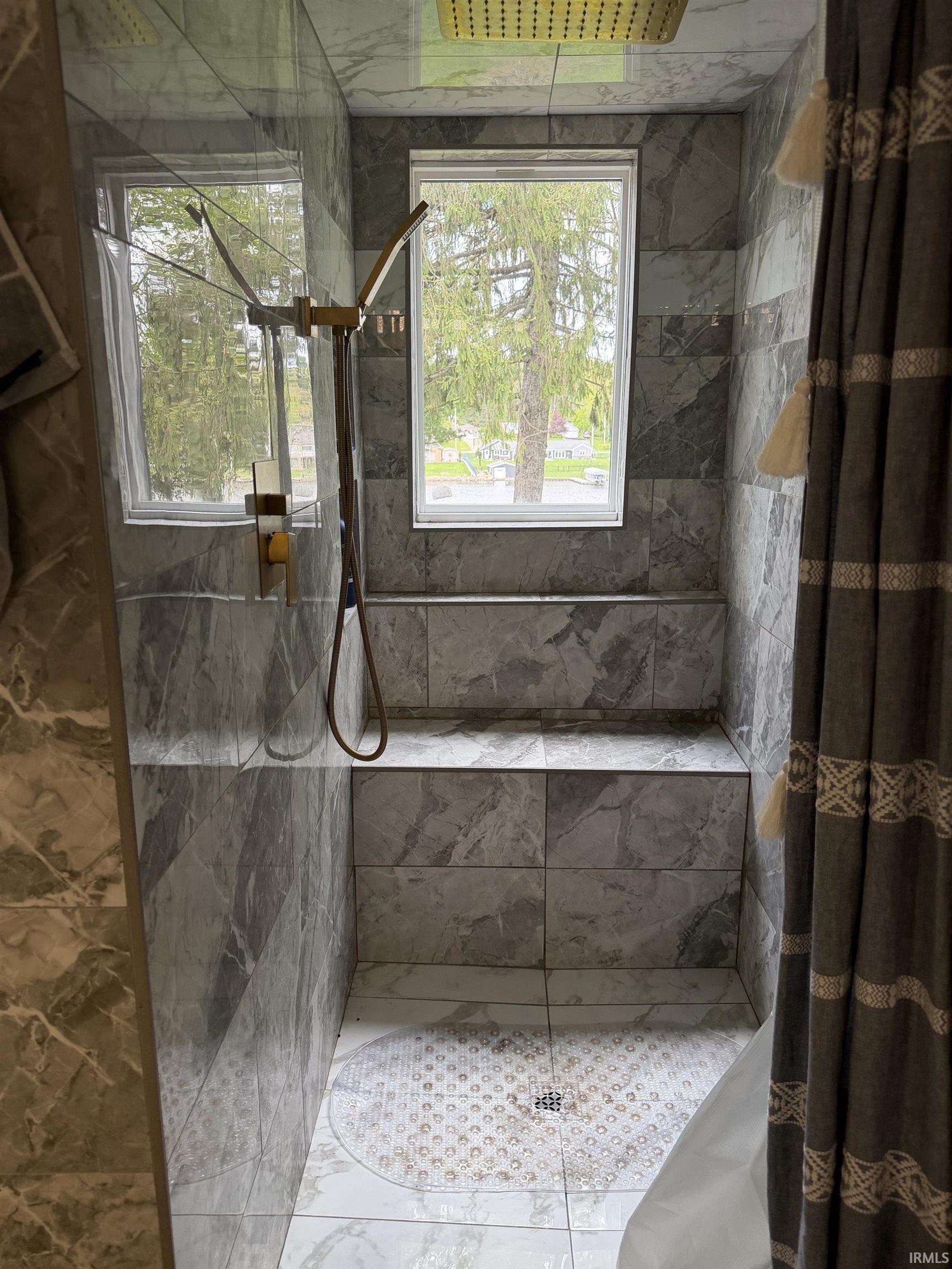 Walk-in shower featuring full-height stone-look tile, built-in bench seating, two windows, and a rainfall showerhead