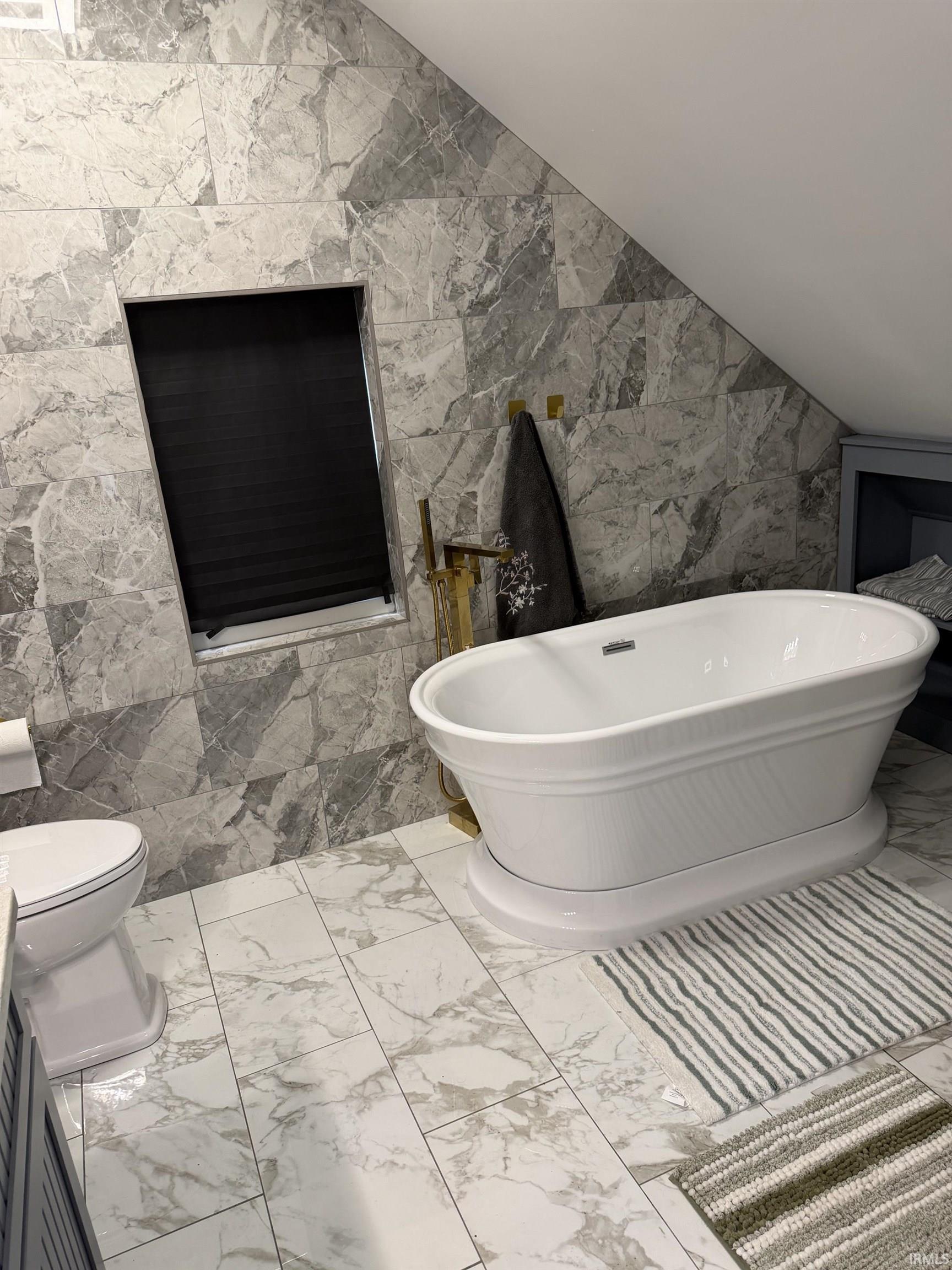 Freestanding soaking tub with floor-mounted brass-finish faucet, accompanied by an in-wall window with a dark privacy shade