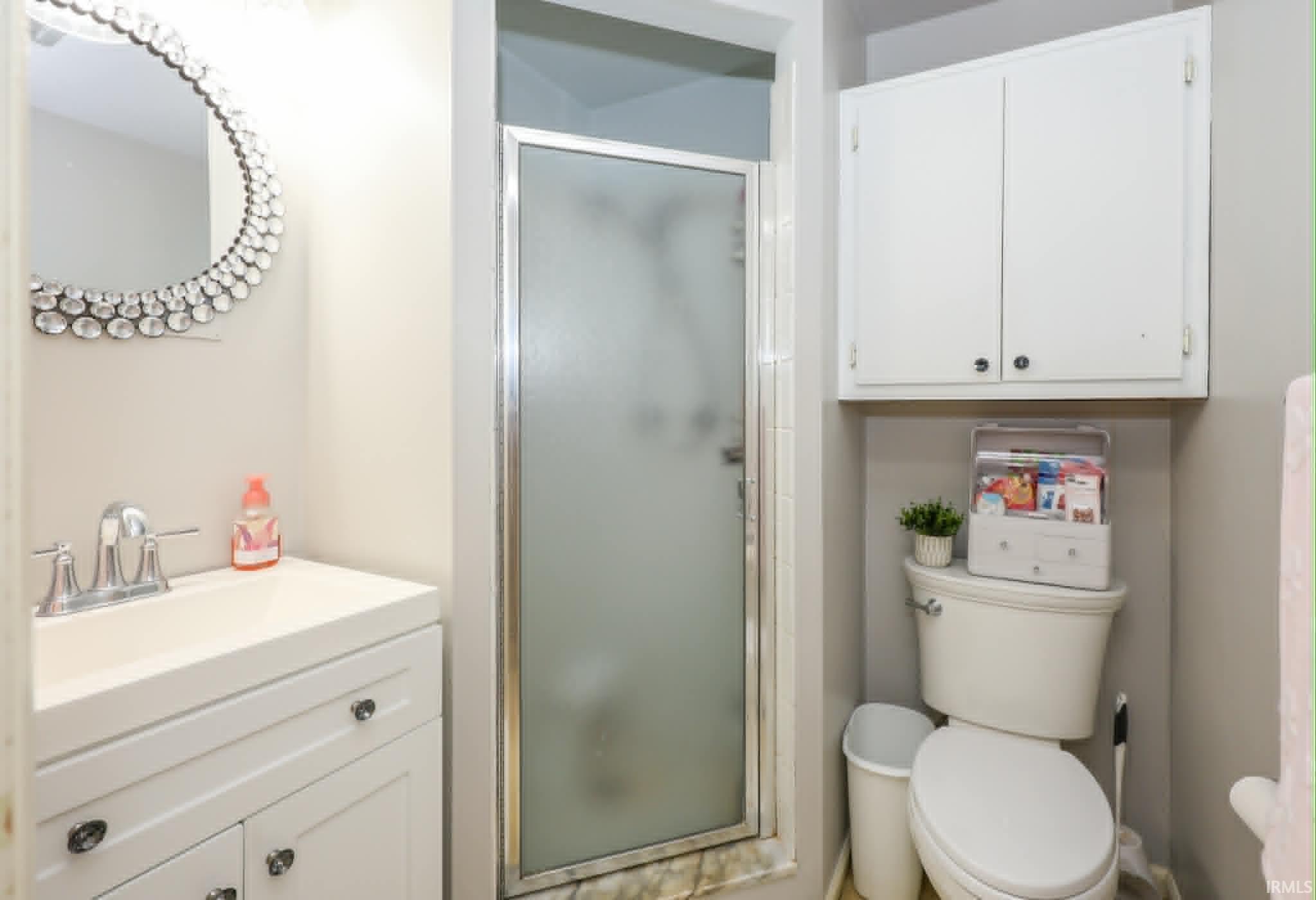 Compact bathroom featuring a shower with a frosted glass door, a vanity with a white countertop and base cabinetry, a circular decorative mirror, and a wall-mounted storage cabinet