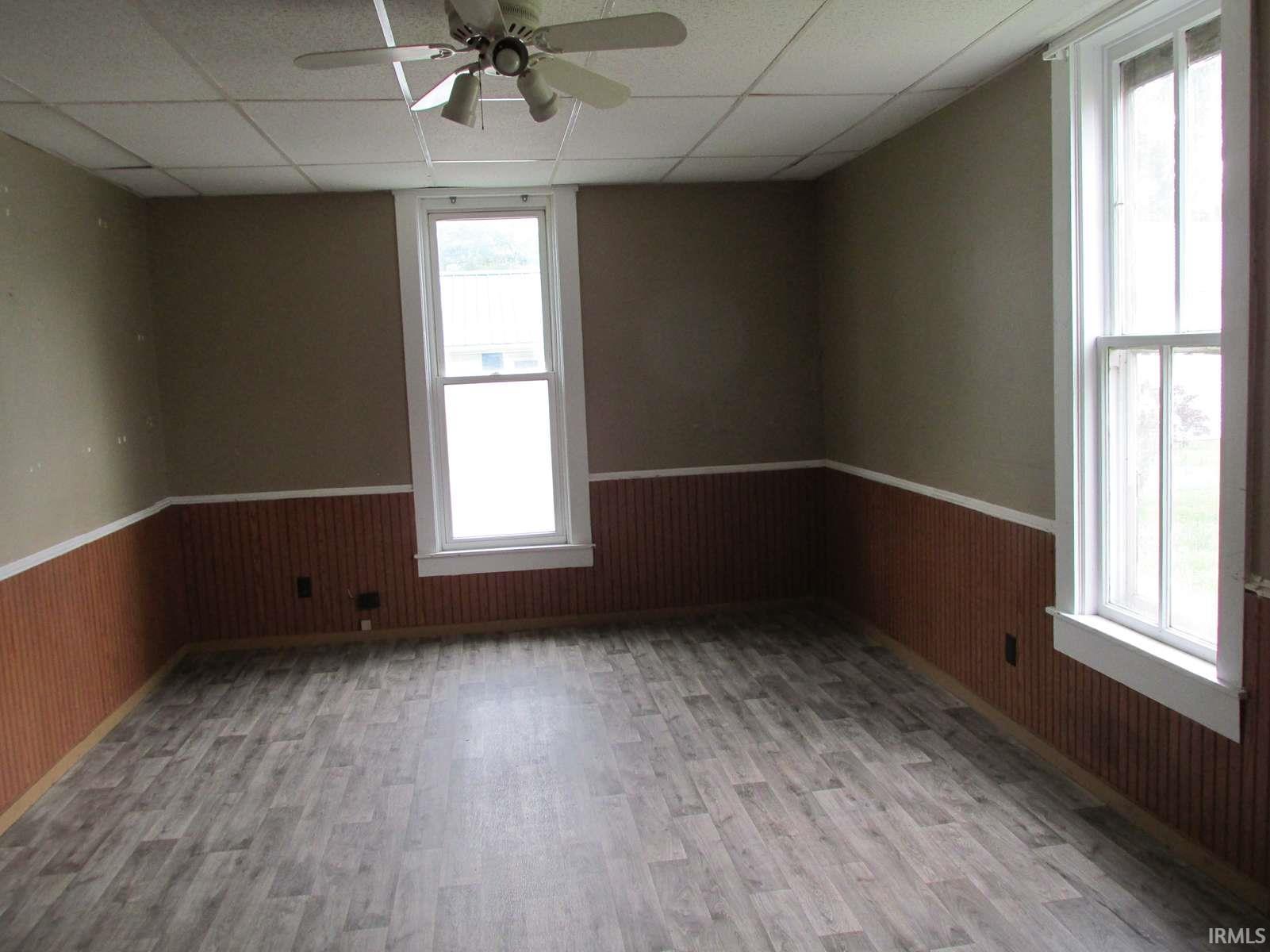 Room featuring light wood-finish flooring, two large windows with white trim, beadboard wainscoting, a drop ceiling, and a ceiling fan with light fixture