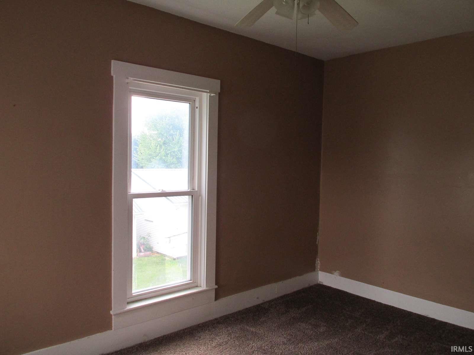 Room featuring a double-hung window with white trim, a ceiling fan with light fixture, brown carpeting, brown painted walls, and white baseboards