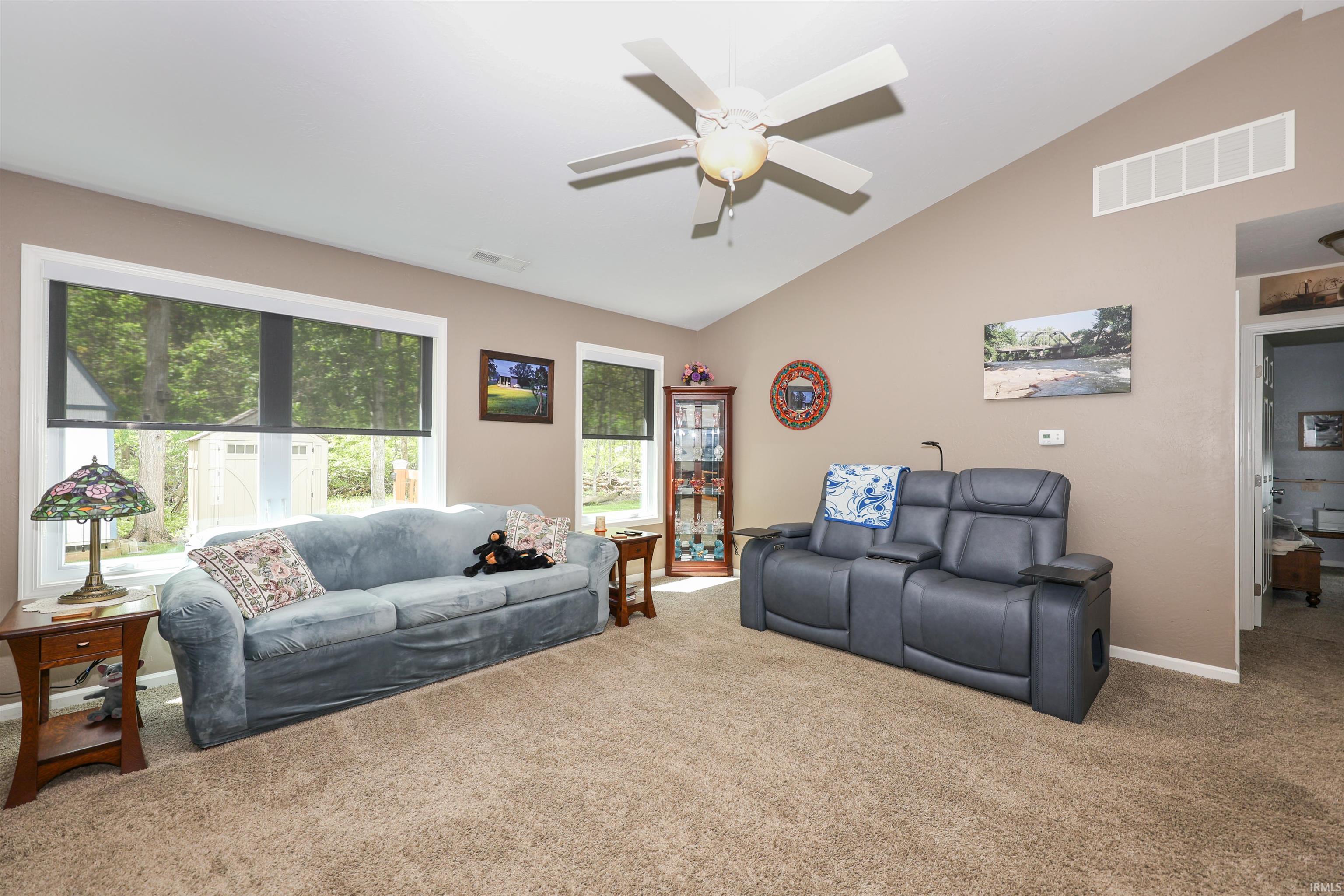 Spacious living area featuring a vaulted ceiling with a ceiling fan, broad windows with roller shades, and plush carpet flooring
