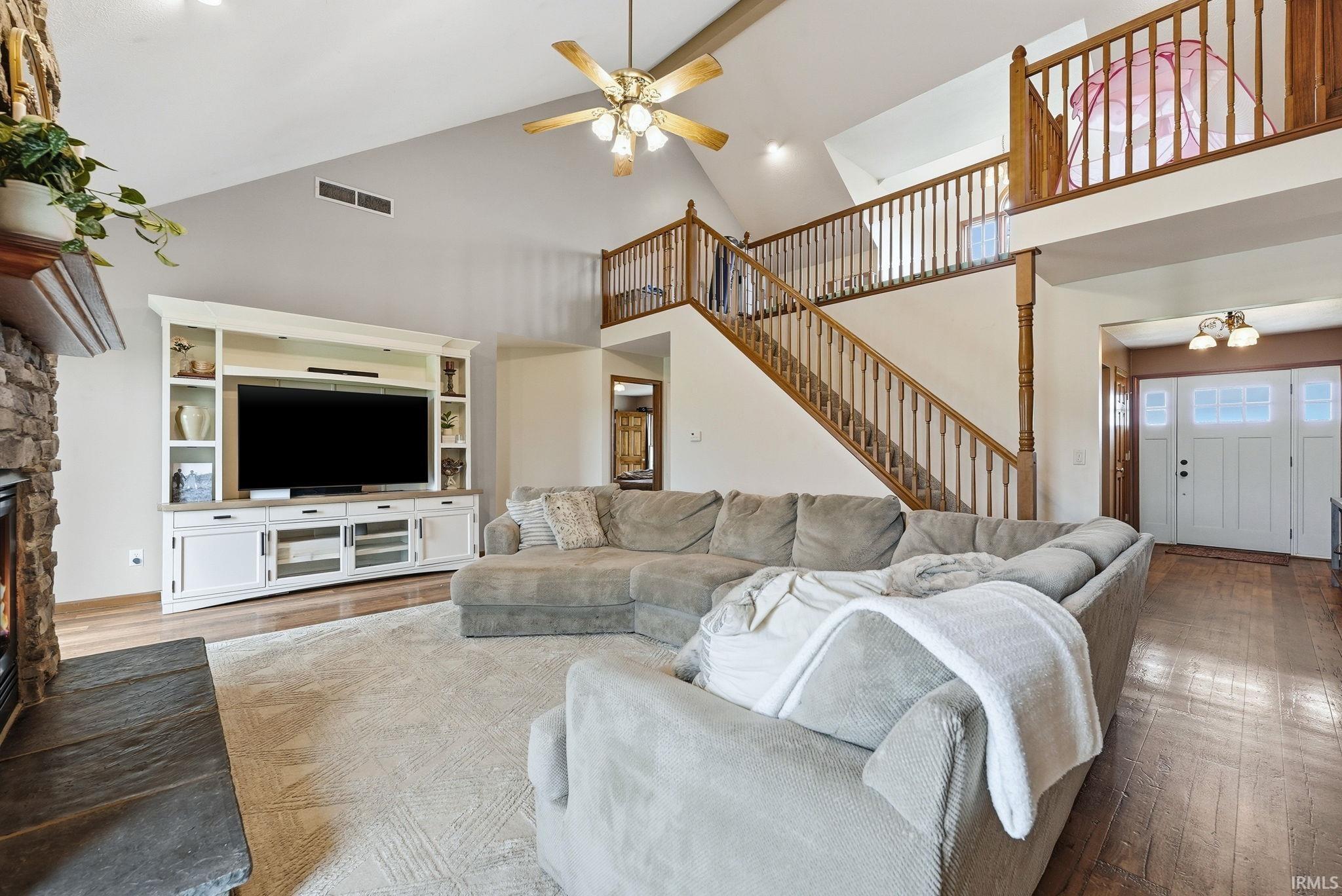 Vaulted ceiling with an overhead fan, stone-faced fireplace with hearth, built-in entertainment center, wood-finish flooring, and a multi-level staircase with wood railings