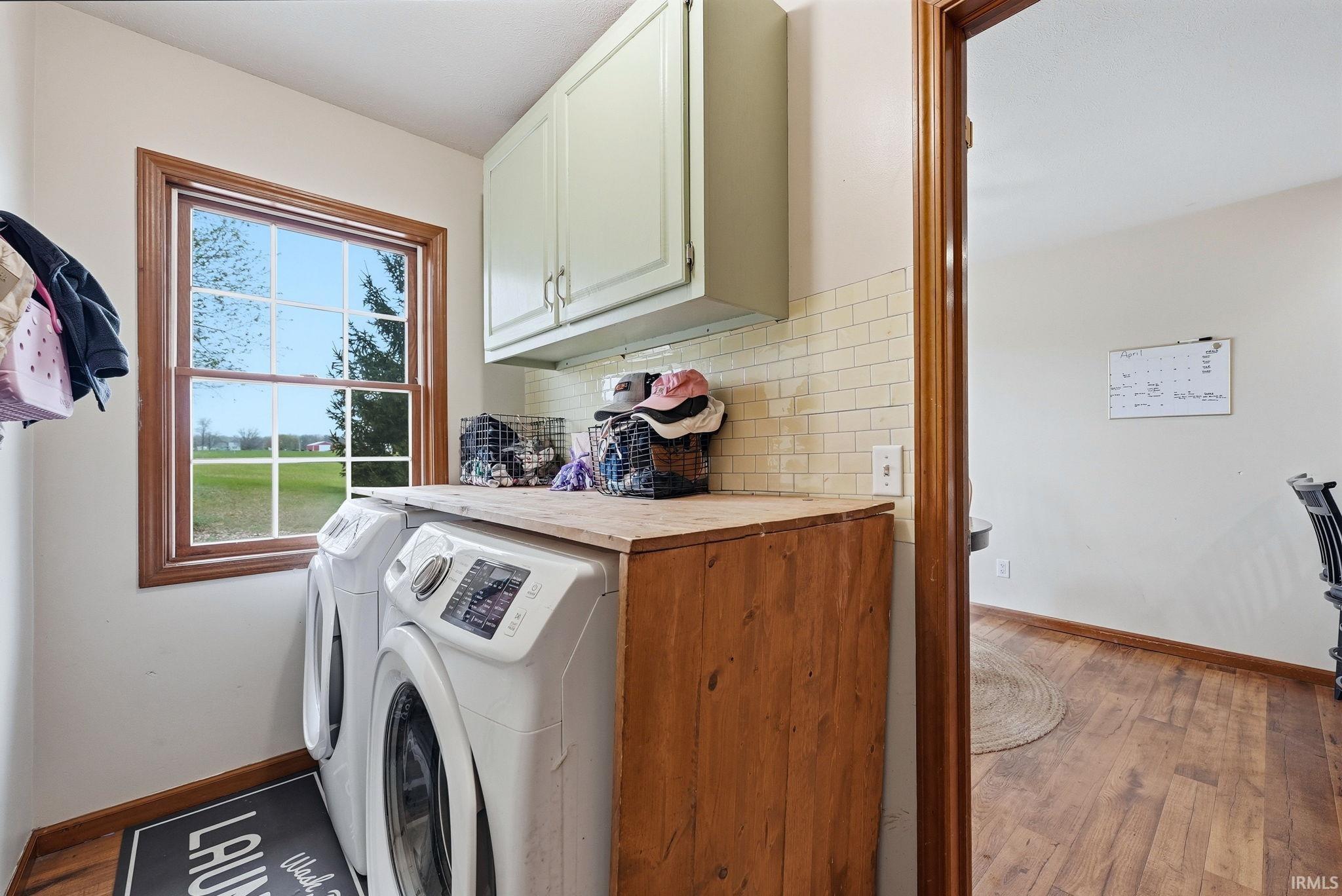 Dedicated laundry space featuring a window, wood-finish flooring, light-toned upper cabinetry, a wood-finish countertop, and a light-toned subway tile backsplash