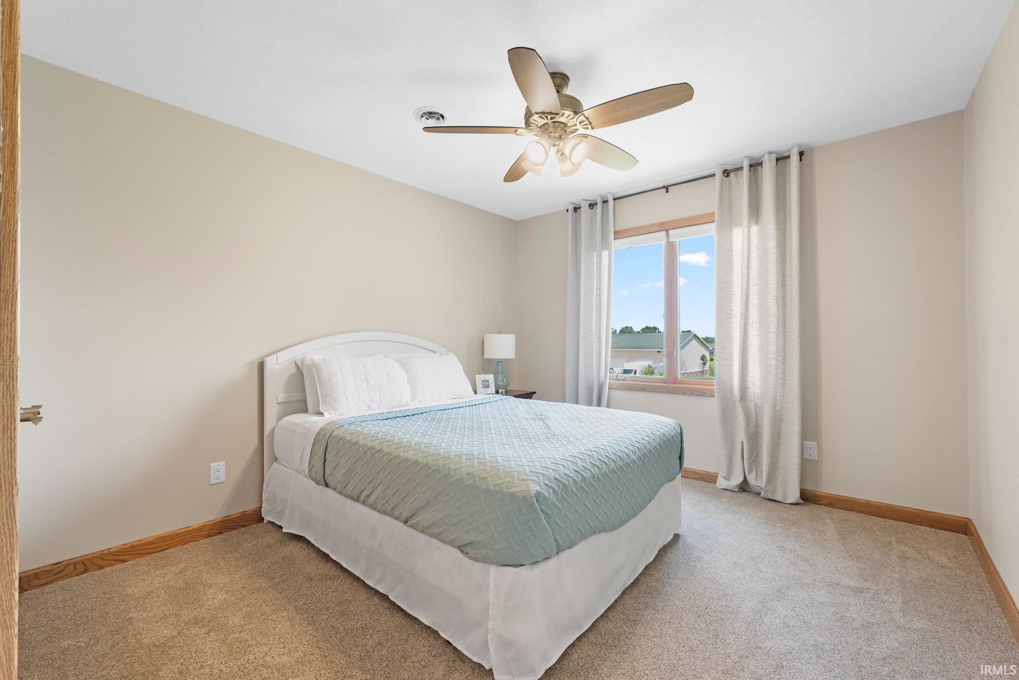 Light-filled room featuring neutral wall tones, plush carpet flooring, a ceiling fan with light fixture, and a window with wood trim