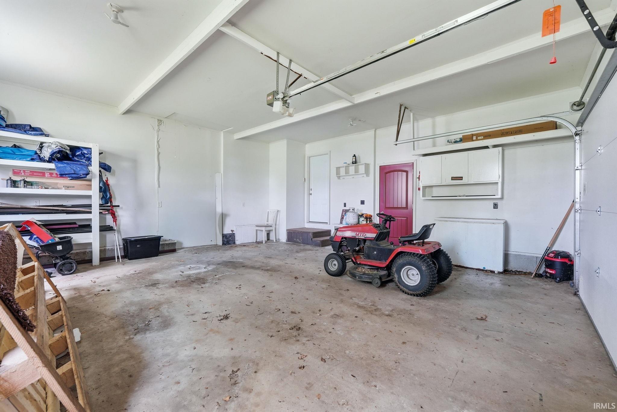 Spacious garage featuring a concrete floor, exposed ceiling beams, built-in shelving, a side access door, and overhead cabinet storage