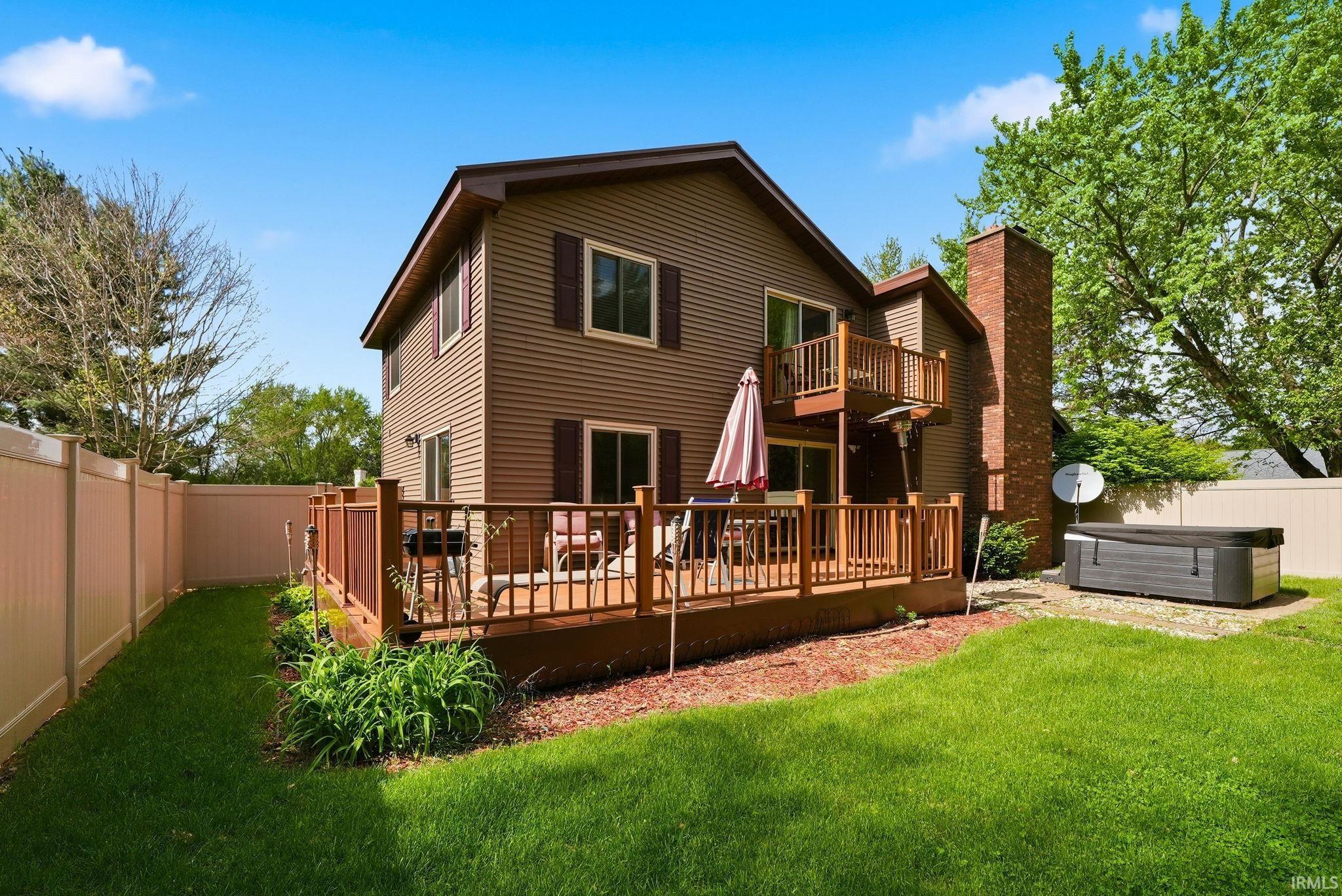 Two-story residential structure featuring horizontal siding, a private second-story balcony, a brick chimney, a ground-level wood deck, and a fenced backyard with a lawn area