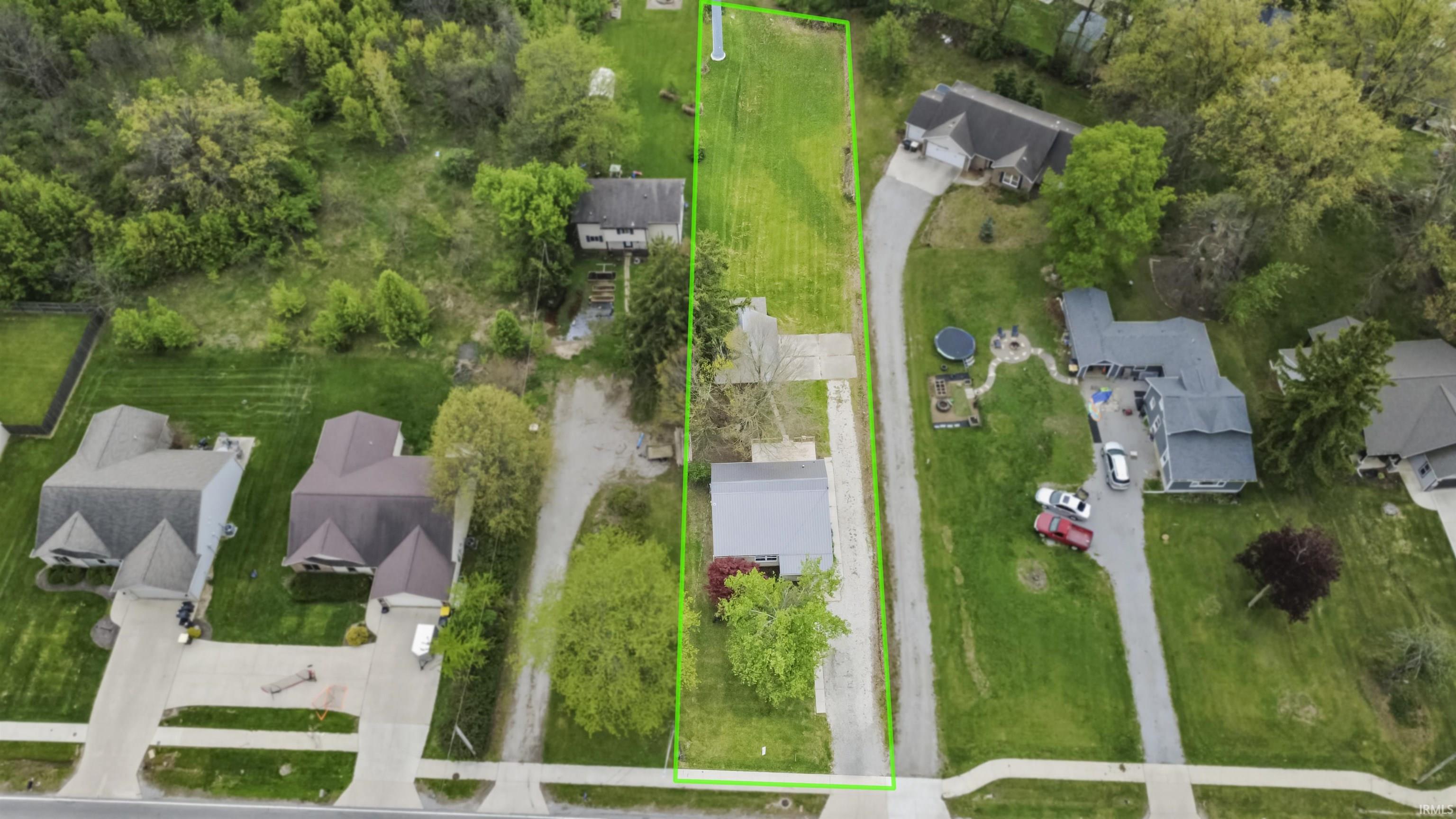 Elongated parcel featuring a substantial grassy area, mature trees, and a gravel driveway accessing a single-story residence with a light gray roof