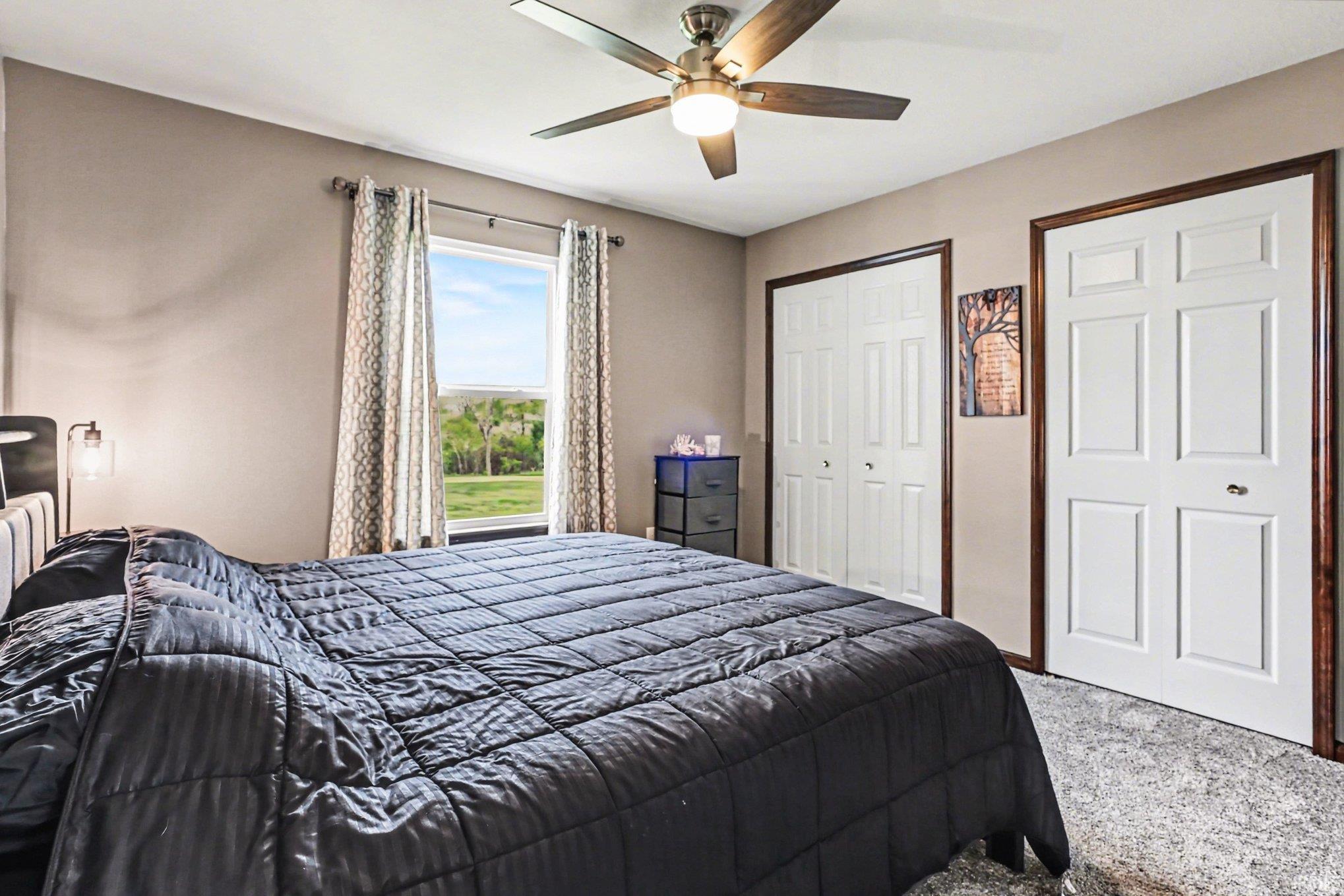 Carpeted bedroom with a window featuring exterior scenery, an overhead ceiling fan with lighting, two sets of bi-fold closet doors, and dark wood trim