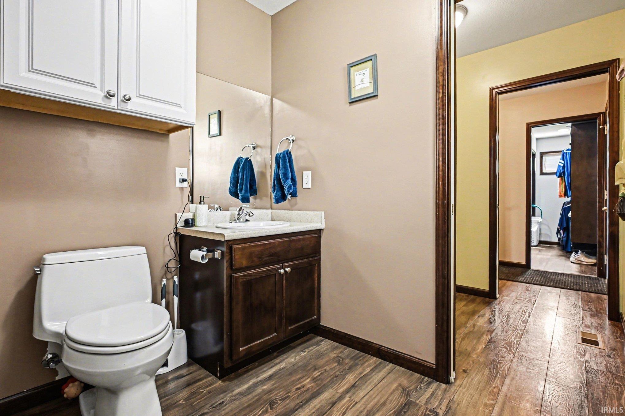Bathroom featuring wood-finish flooring, a white toilet, a vanity with a white countertop and dark wood-finish cabinetry, and white wall-mounted storage cabinets