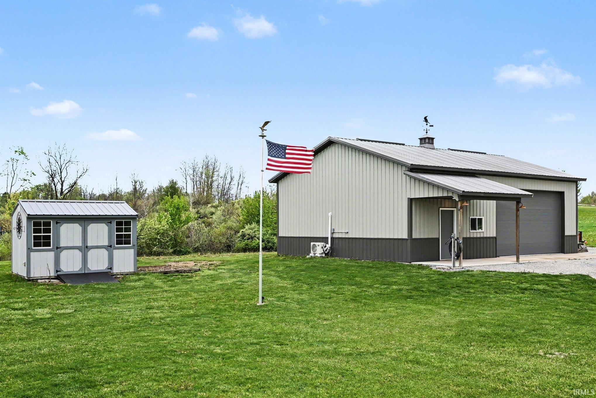 Expansive green lawn featuring a metal-sided outbuilding with a porch entry and two garage doors, alongside a separate storage shed with double doors and window accents