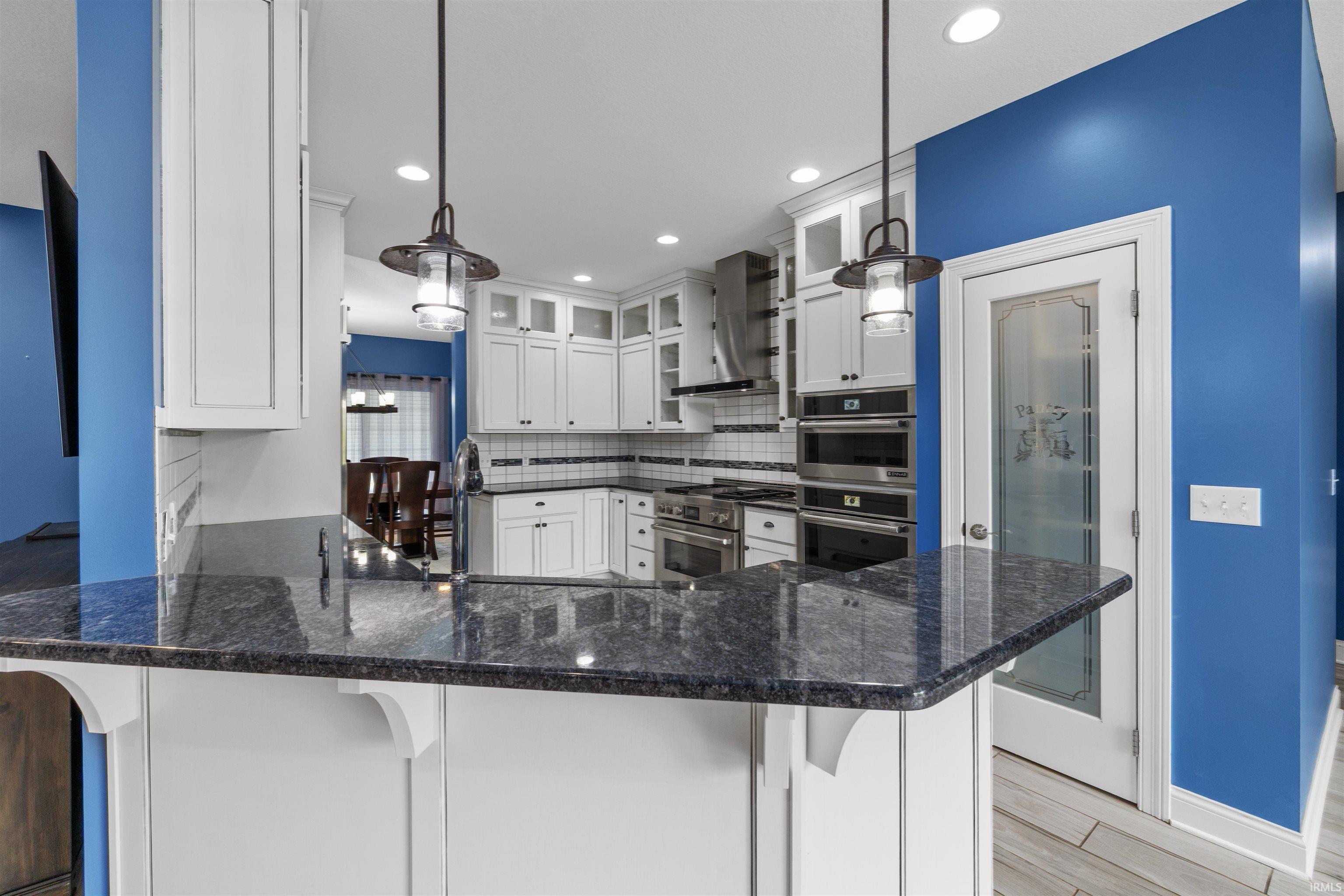 Kitchen featuring a granite-finish countertop peninsula, white cabinetry with glass-front accents, stainless steel appliances, pendant lighting, and a pantry with a frosted glass door