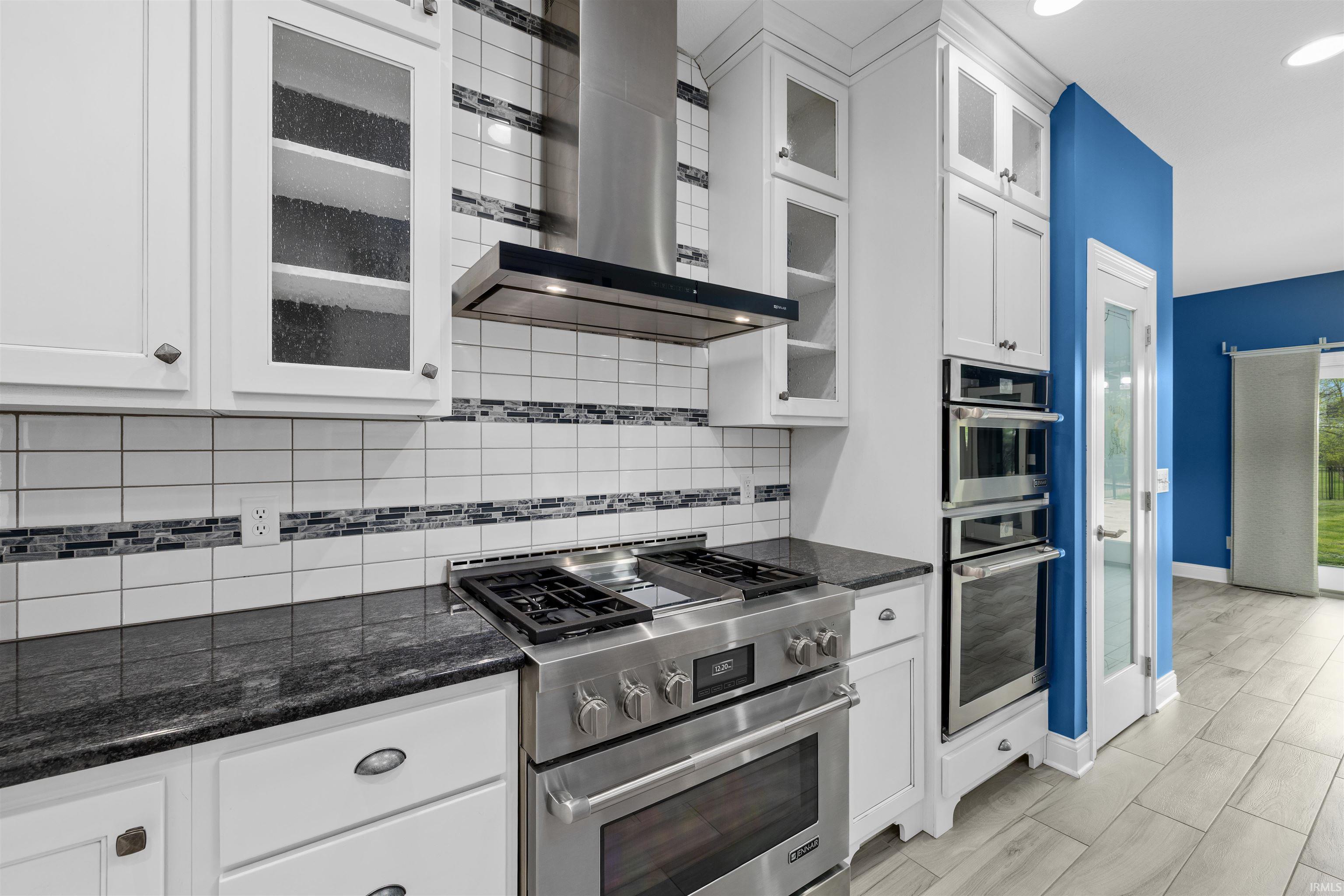 Gourmet kitchen featuring stainless steel appliances, granite countertops, white cabinetry, a subway tile backsplash with decorative accents, and wood-finish flooring