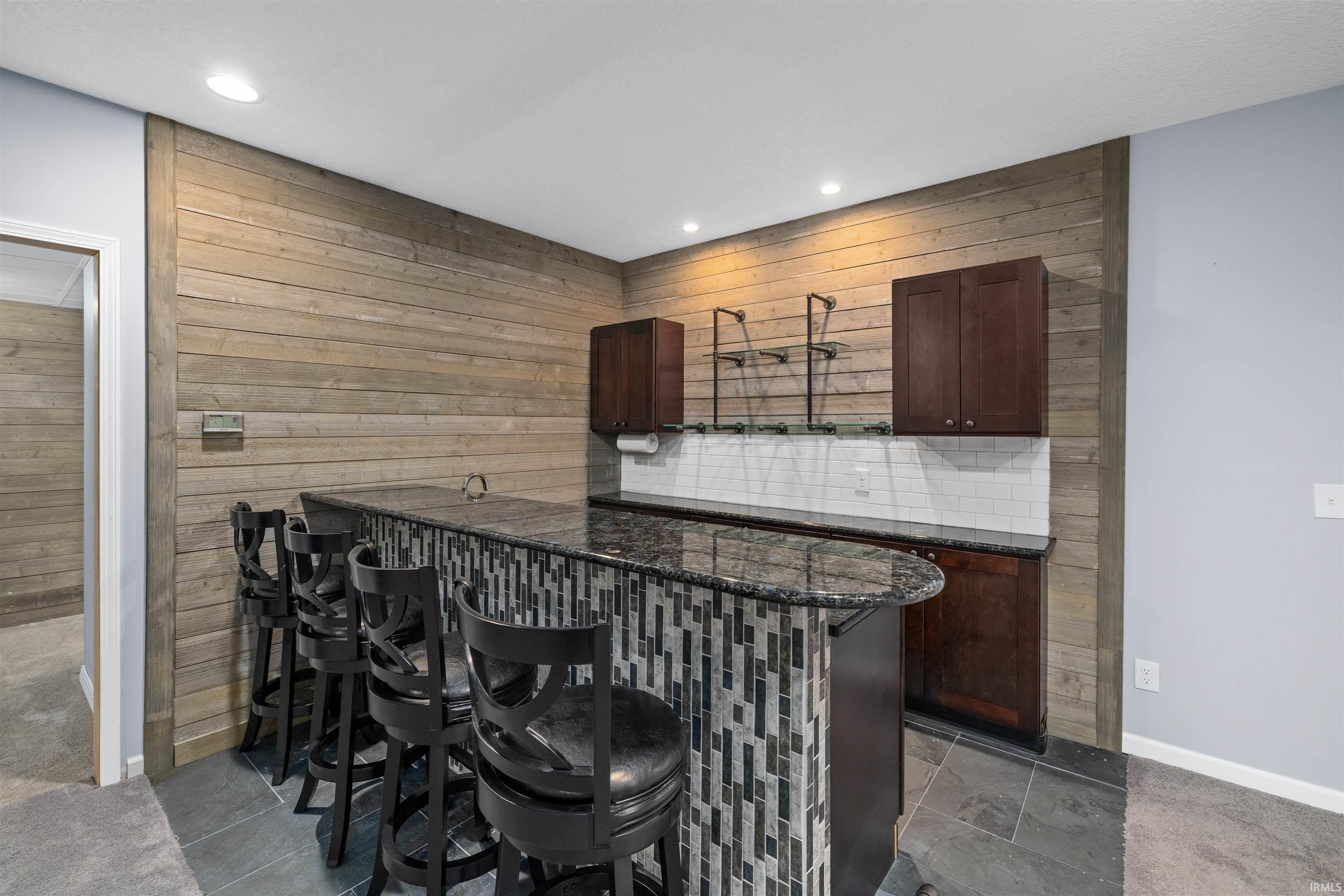 Wet bar featuring a granite countertop, mosaic tile bar facing, wood-finish accent wall, dark wood cabinetry, and a white subway tile backsplash