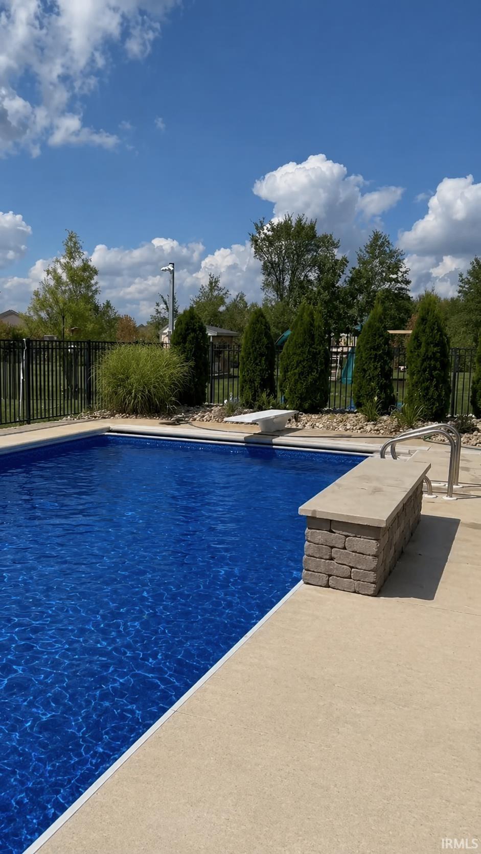 In-ground swimming pool featuring a diving board, surrounding concrete patio, and dark metal fencing