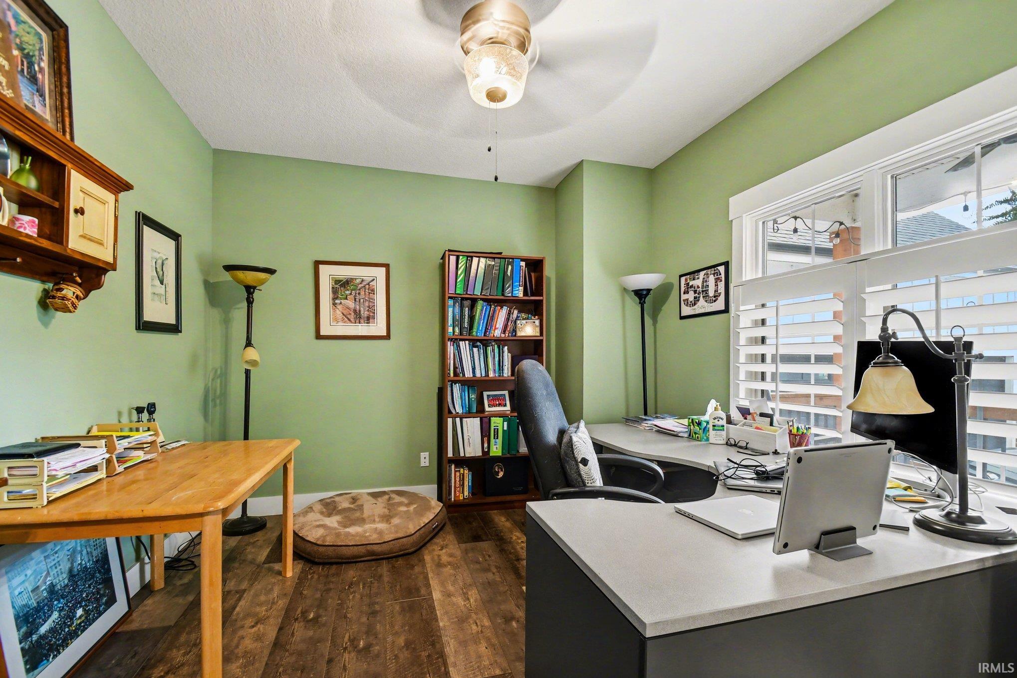 Dedicated workspace featuring wood-finish flooring, two large windows with white louvered shutters, a built-in bookshelf, and a ceiling fan with integrated lighting