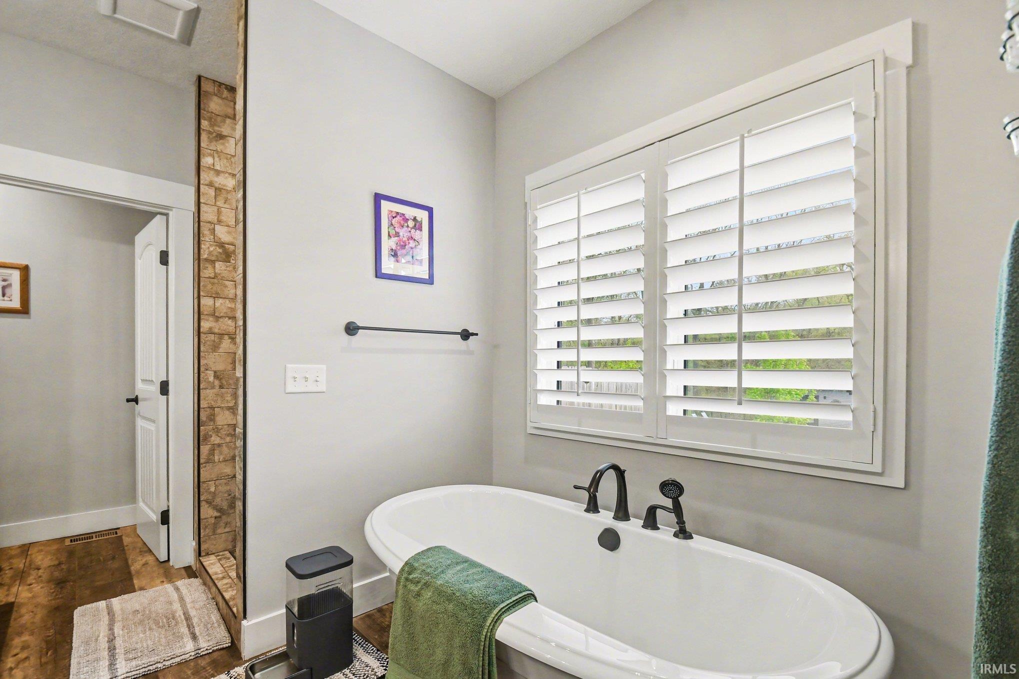 Freestanding soaking tub with dark bronze fixtures, framed by a window with white plantation shutters