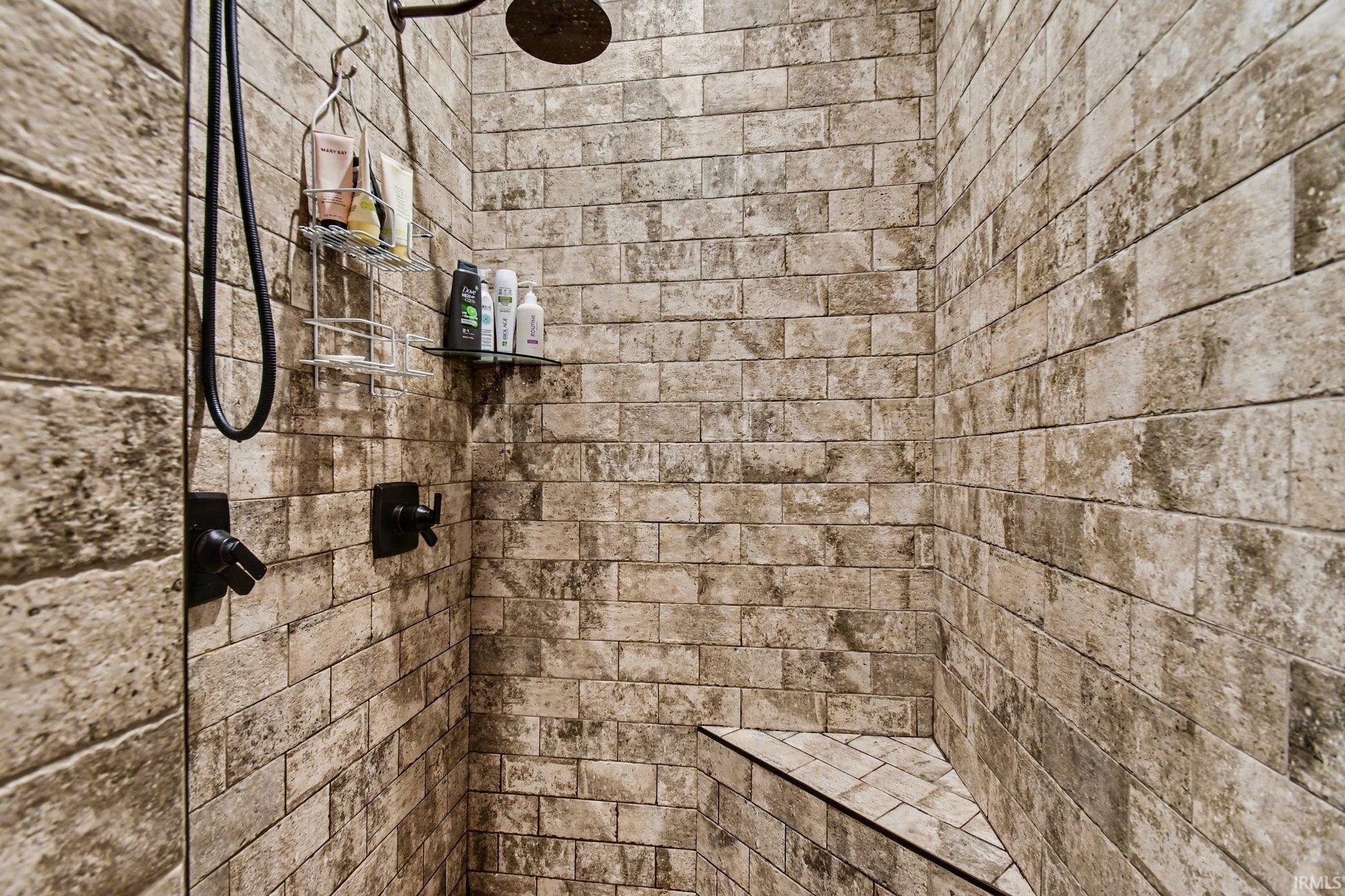 Walk-in shower featuring textured rectangular tile walls, a built-in corner bench, a rainfall showerhead, and matte black fixtures